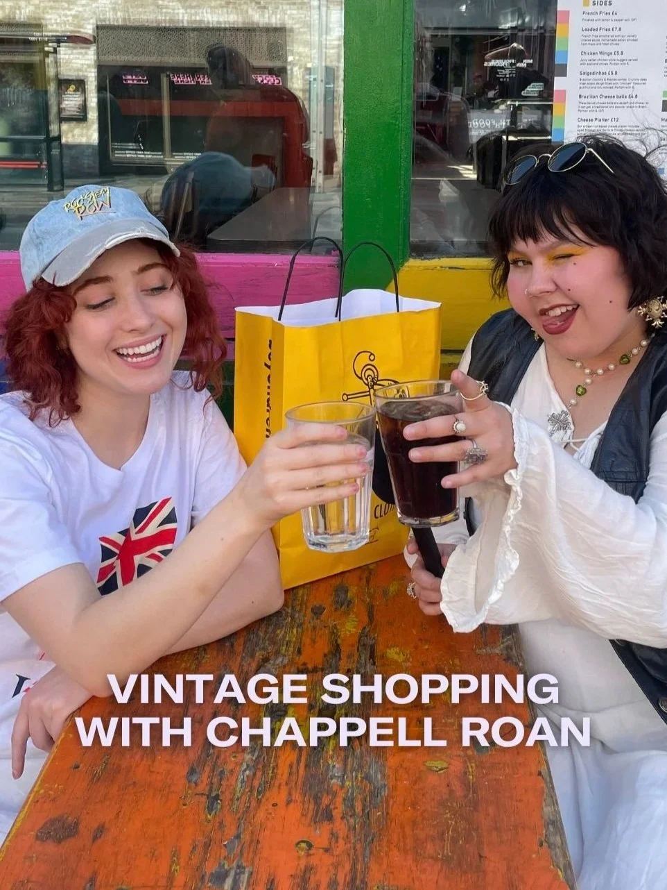 Two women sitting at a rustic wooden table outside a colorful shop, toasting with glasses of dark soda, smiling, with shopping bags nearby, celebrating a vintage shopping trip.