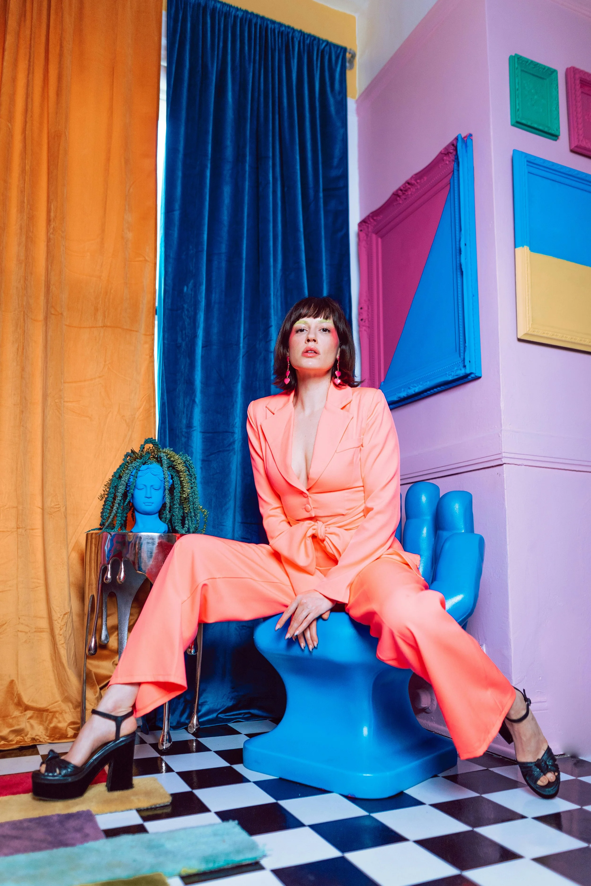 Woman in a coral jumpsuit sitting on a blue toilet in a colorful room with yellow, pink, and blue walls, black and white checkered floor, and abstract art on the walls.