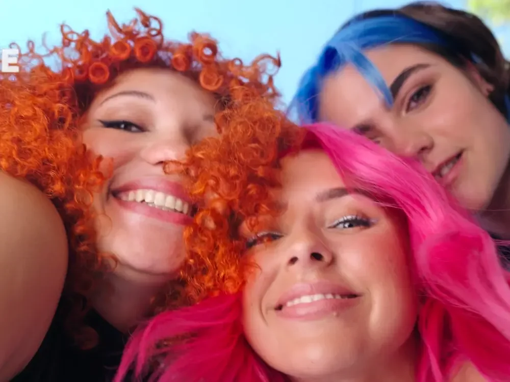 Three women smiling closely together with vibrant hair colors: one with bright orange curly hair, another with hot pink wavy hair, and the third with a mix of dark brown and blue hair.