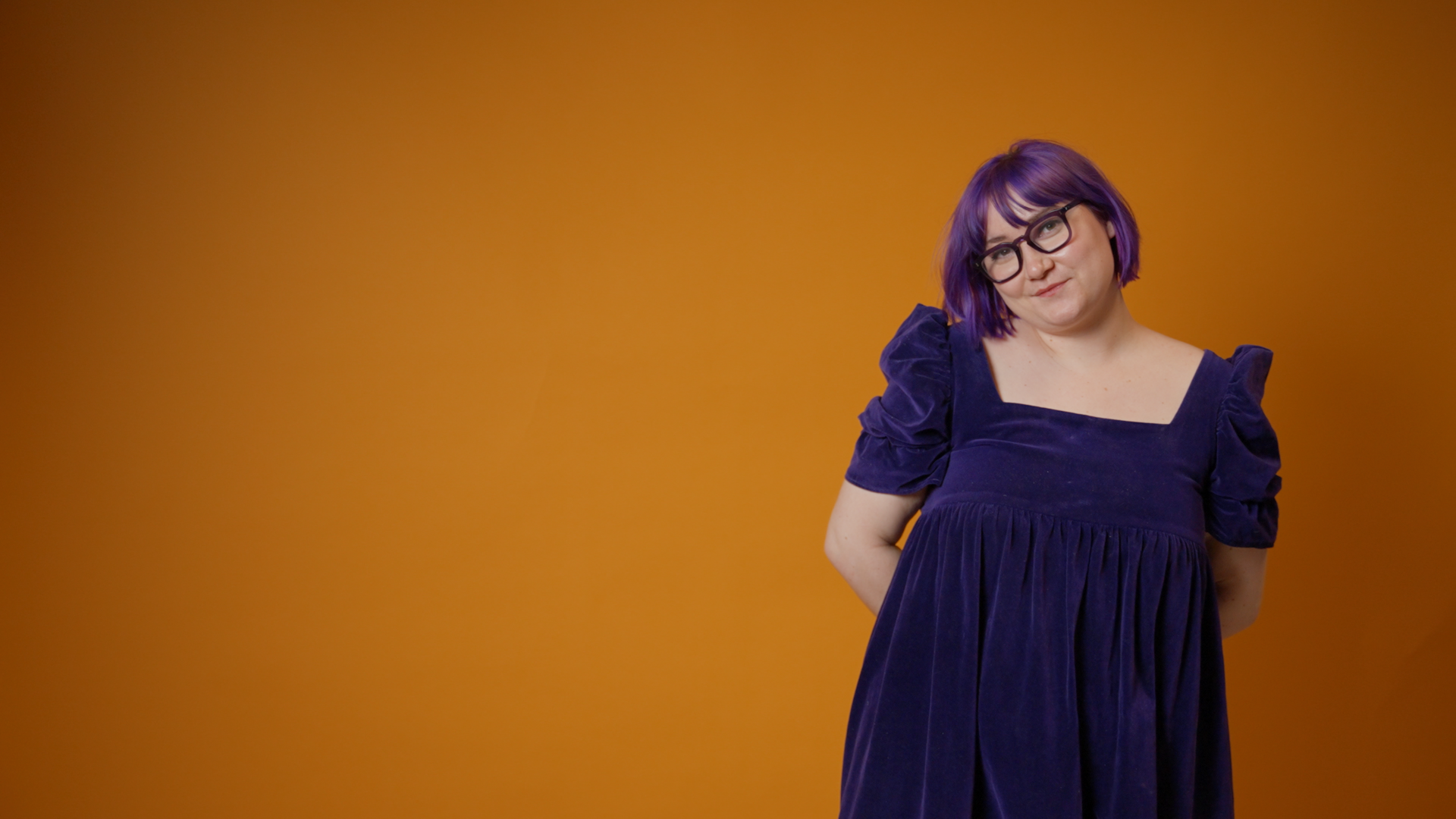 Woman with purple hair and glasses in a dark purple dress standing against an orange background.