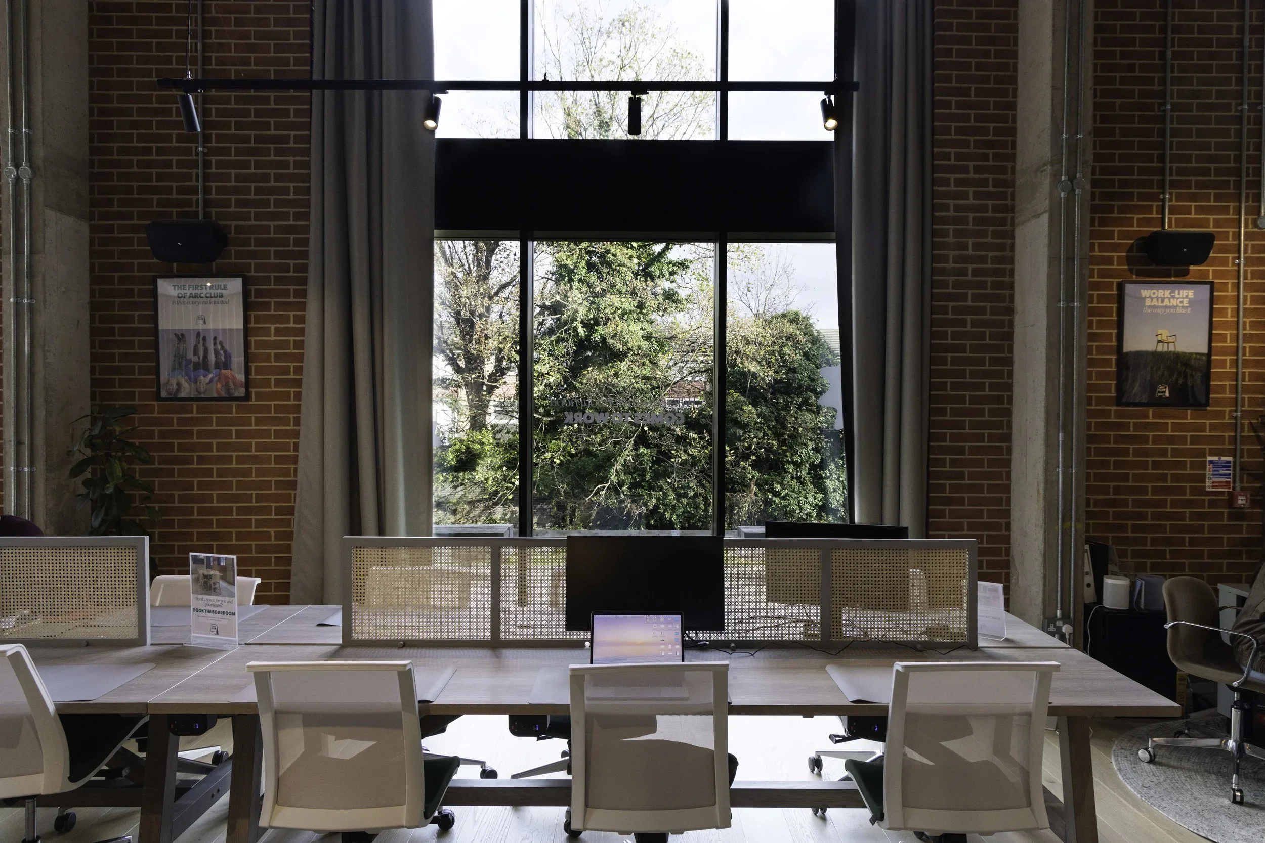 Modern office workspace with a large window showing trees outside, brick walls, a long desk with multiple white ergonomic chairs, computer monitors, and decorative posters on the brick walls.