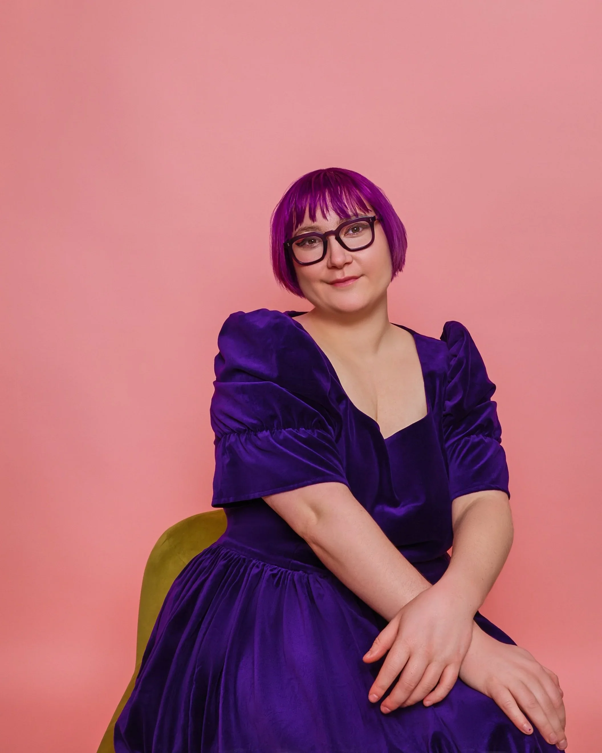 Woman with purple hair and glasses sitting on a green chair against a pink background, wearing a purple velvet dress.