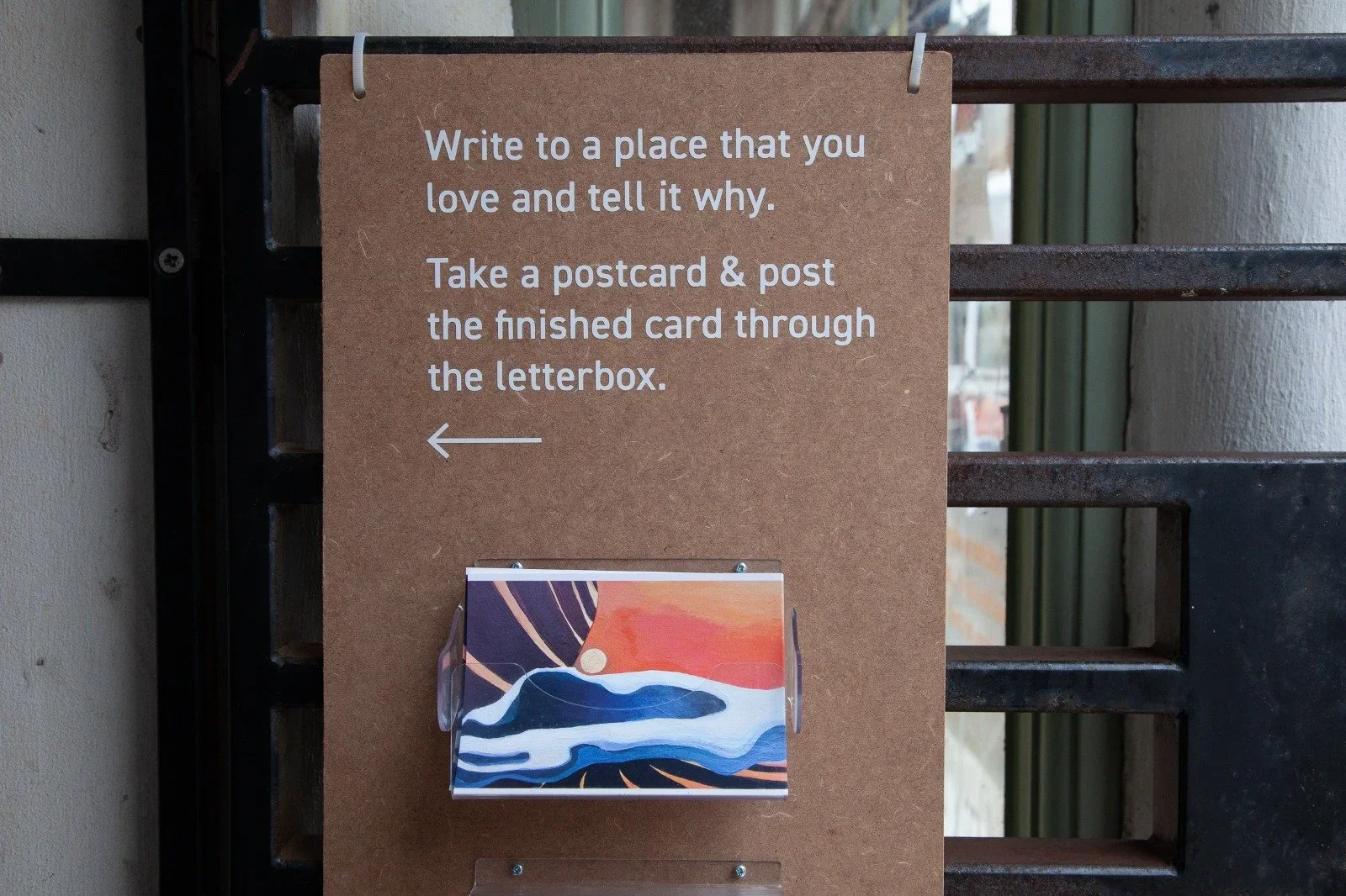 A brown sign with white text and an arrow pointing left. Below the sign is a colorful postcard in a clear plastic holder. The background features a black metal gate and a beige wall.