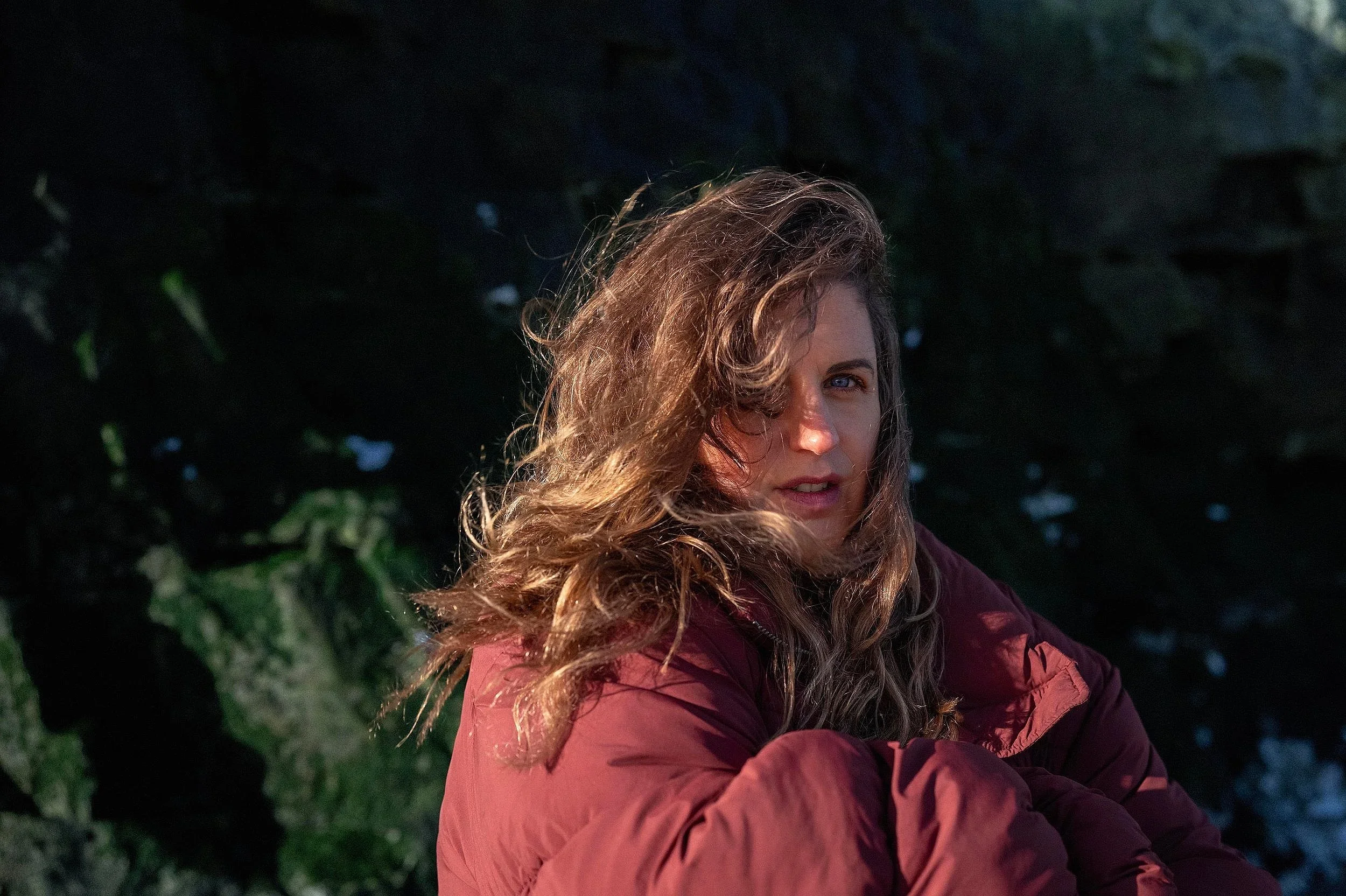 A woman with wavy, light brown hair, wearing a maroon jacket, looks at the camera with a slight smile, outdoors during dawn or dusk with dark rocks and greenery in the background.