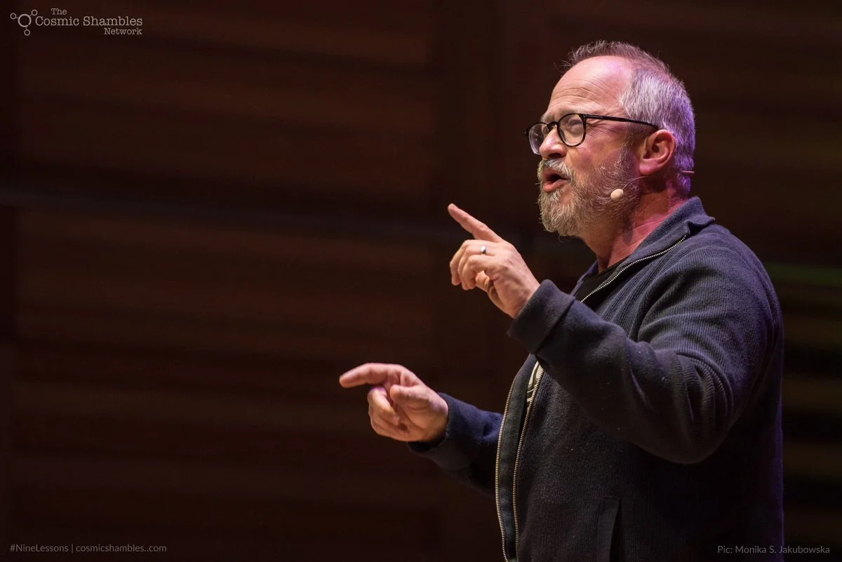 A middle-aged man with glasses, a beard, and gray hair is speaking on stage. He is wearing a dark zip-up sweater and a microphone headset, and is gesturing with his right hand while pointing with his left hand. The background is dark with colored lights.