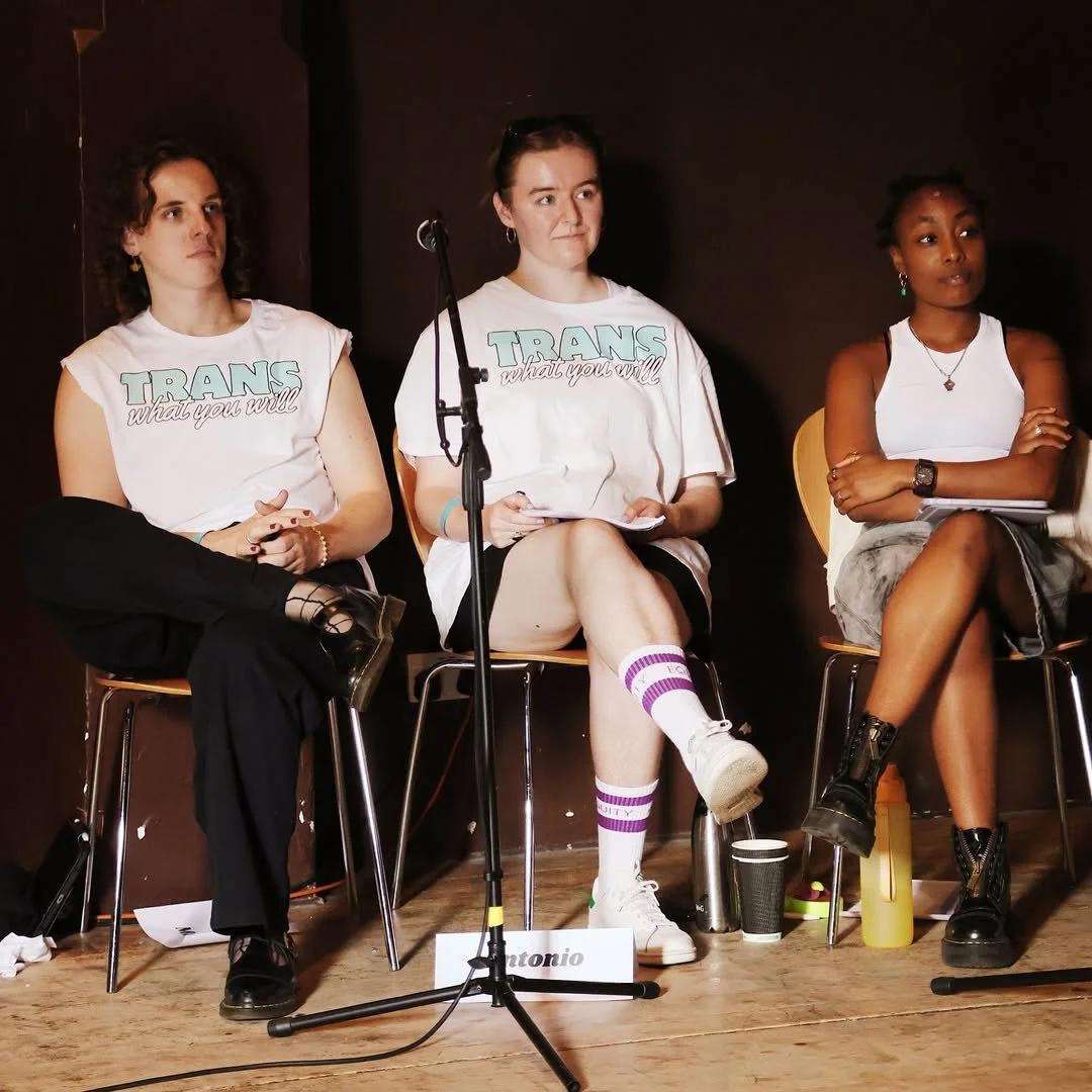 Three women sitting on chairs during a panel discussion, two women are wearing white t-shirts with the phrase 'TRANS what you will' printed on them.