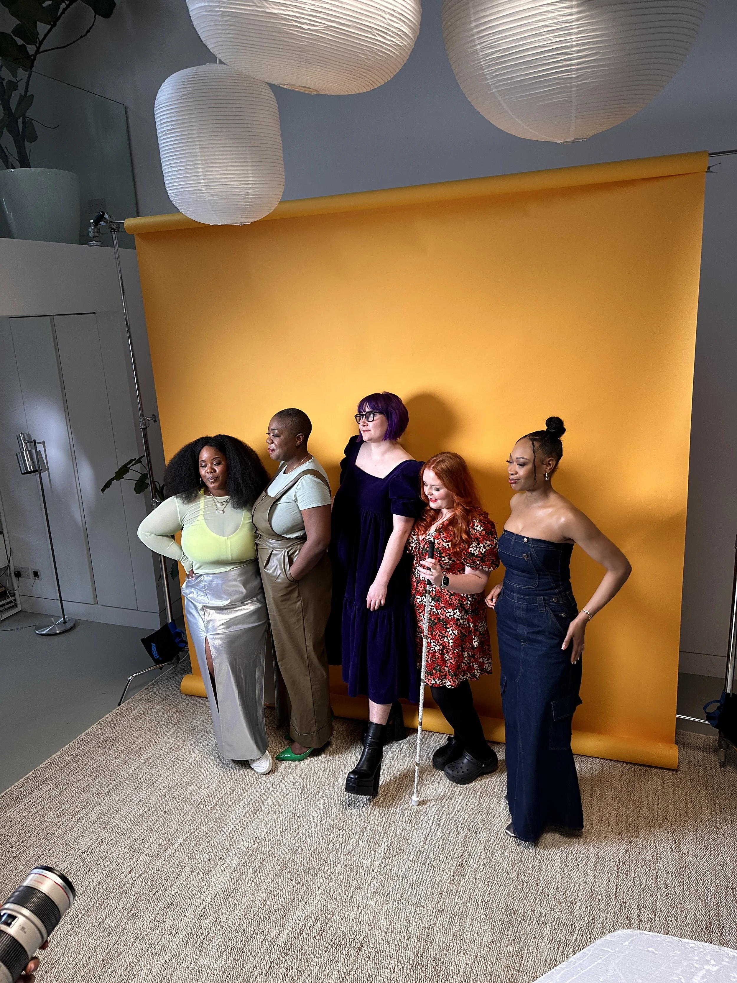 Five women posing in front of a yellow backdrop in a photography studio, with paper lanterns hanging from the ceiling.