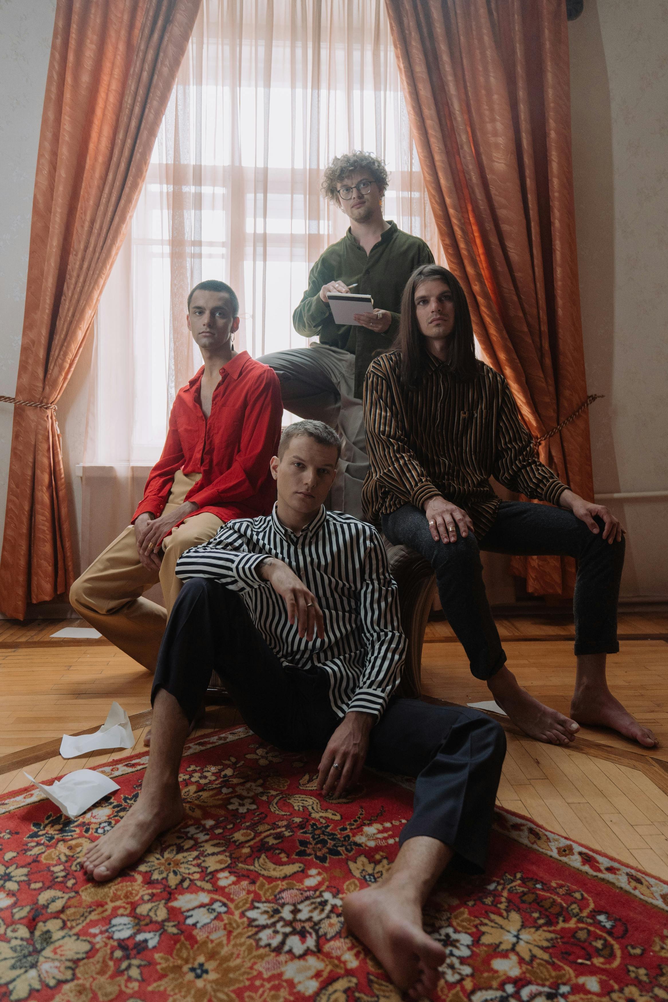 Five young men posing inside a room with orange curtains and a patterned rug. One is sitting on the floor with legs crossed, another is sitting on an armrest, two are seated on a chair, and one is standing behind them, holding a notepad.