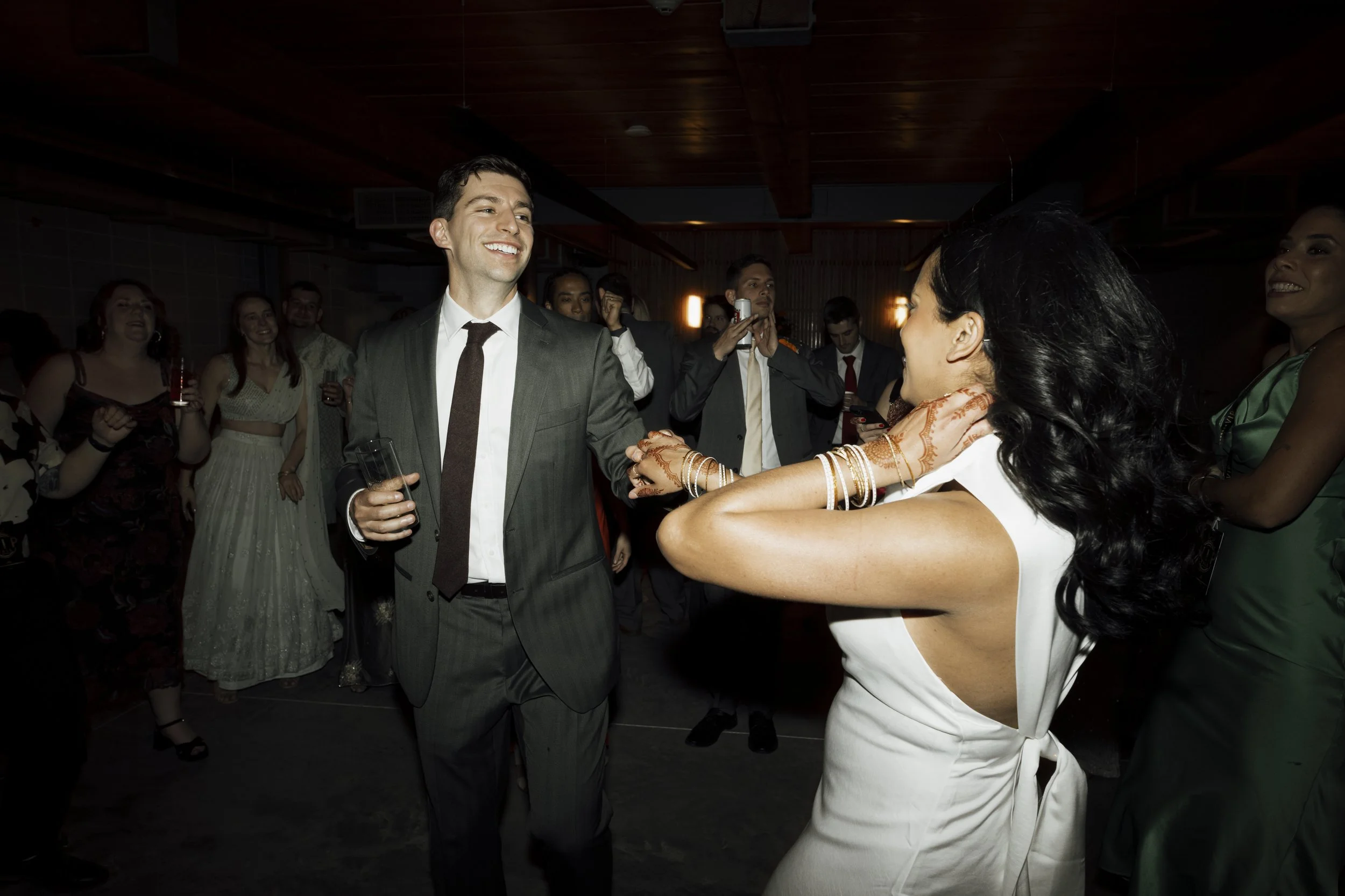 A groom in a gray suit and a bride in a white dress are dancing and holding hands at an upstate NY wedding with other guests in the background.