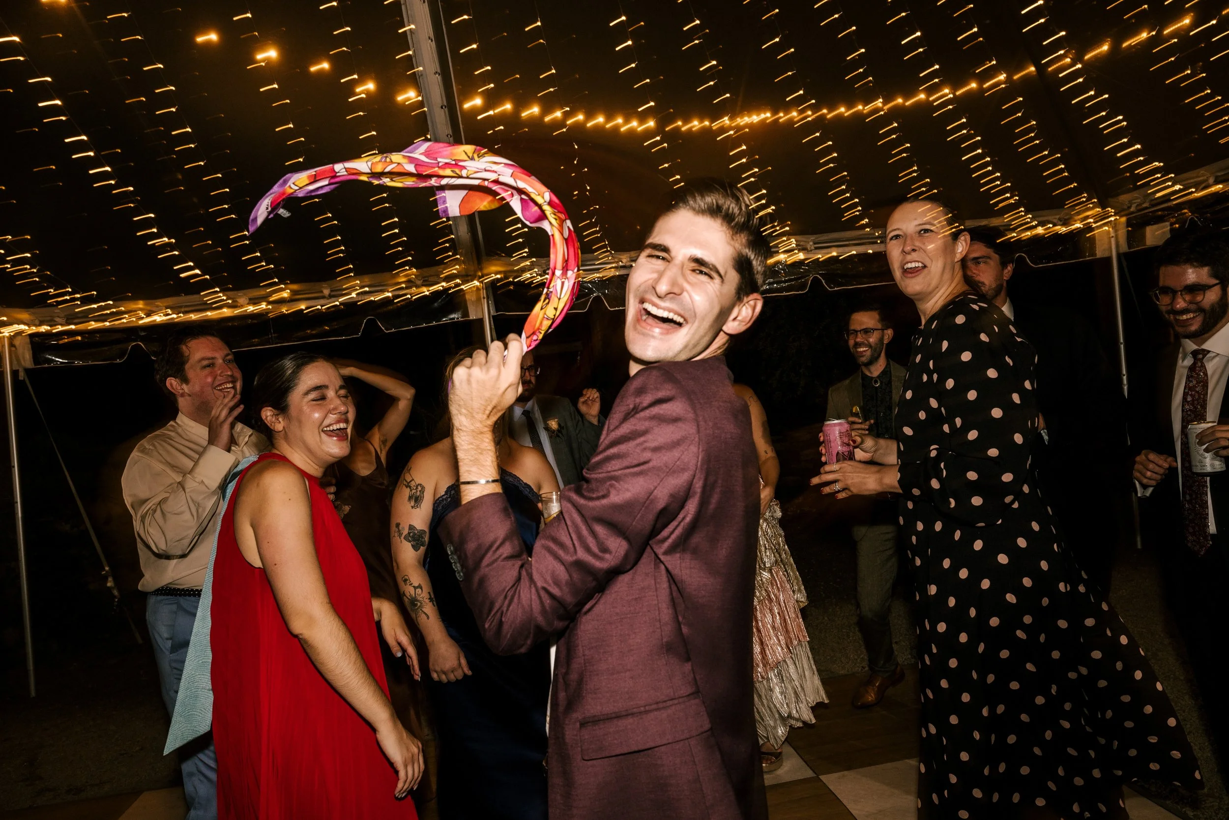 People celebrating and dancing under string lights at an upstate NY wedding, with a man holding a colorful umbrella.