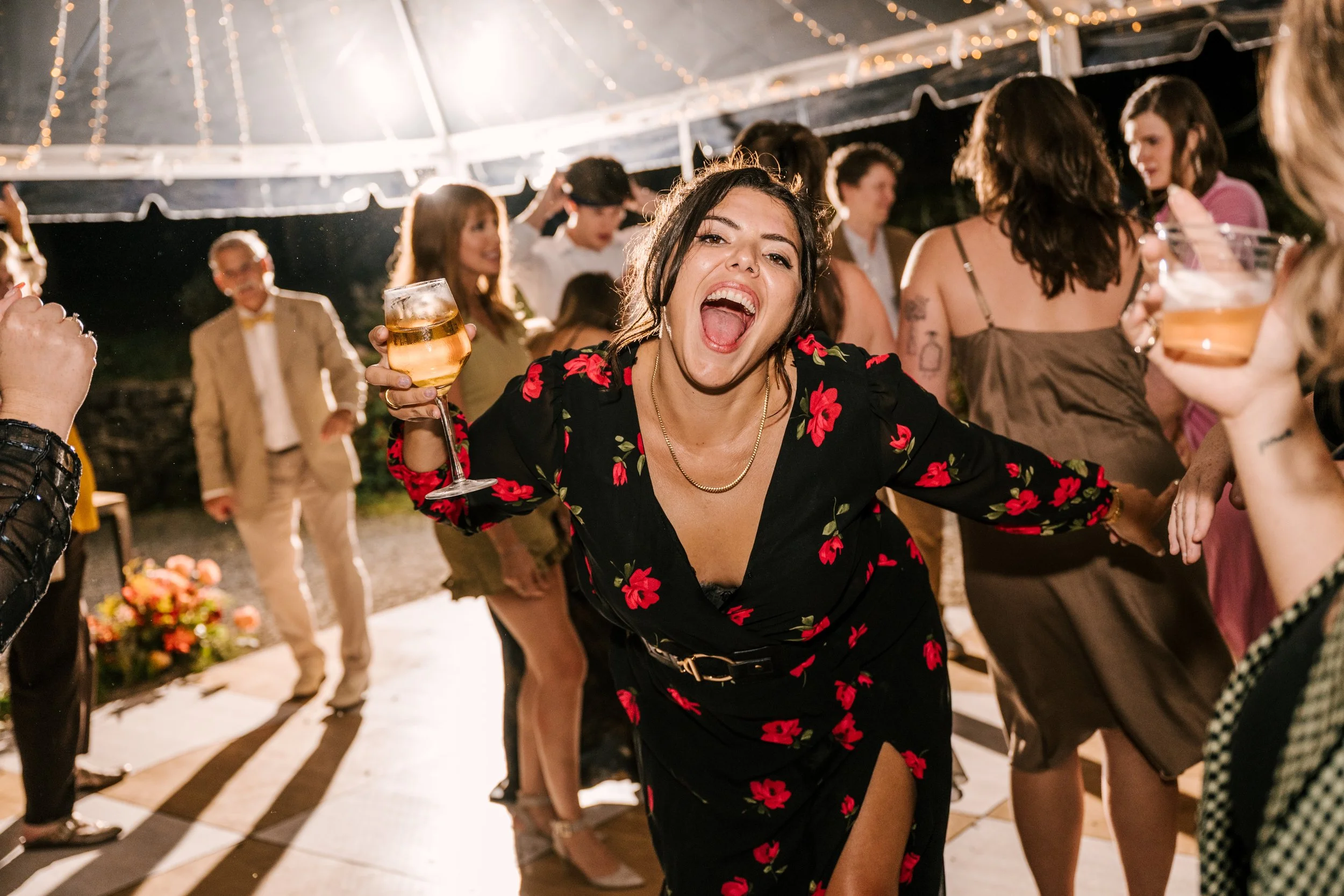 A woman in a black dress with red floral pattern dancing and smiling with a glass of wine at an upstate NY wedding under a large tent with string lights.