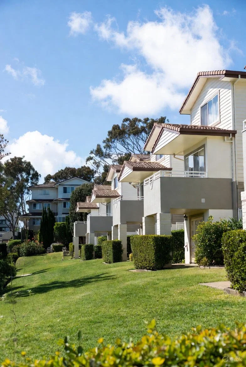 Eden Grounds and Gardens Central Coast Commercial and Strata garden care and green waste removal. A row of modern white residential buildings with balconies, surrounded by green lawns, hedges, and trees under a partly cloudy sky.