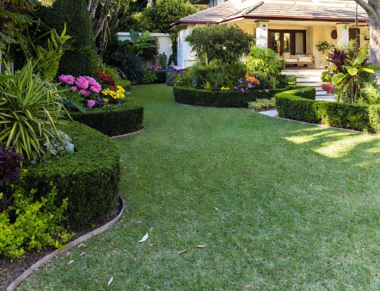 Well-maintained backyard garden with green grass, colorful flower beds, and landscaped bushes surrounding a house patio with seating.