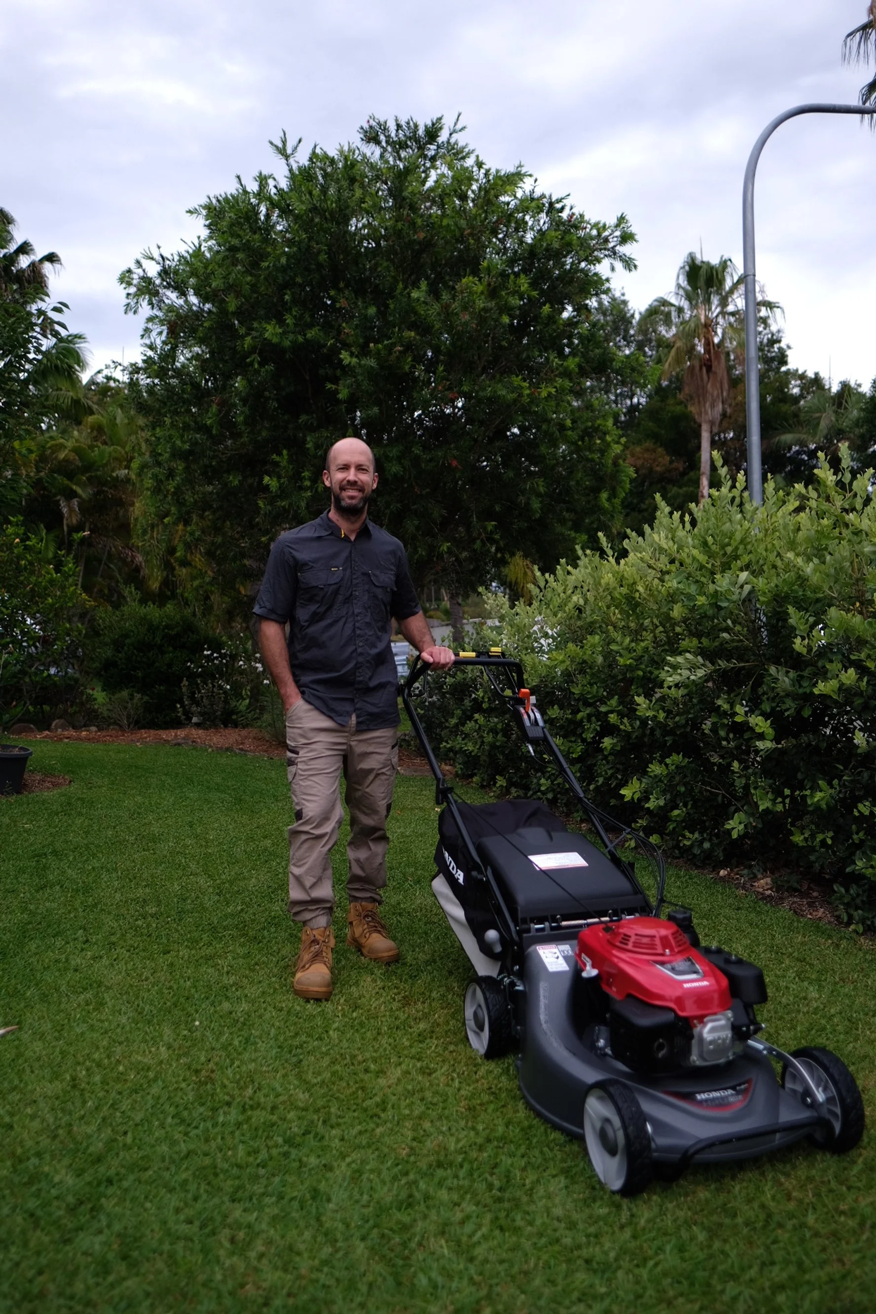 A man in outdoor attire standing on a well-maintained lawn, holding a red and black lawn mower with a natural background of trees and bushes.