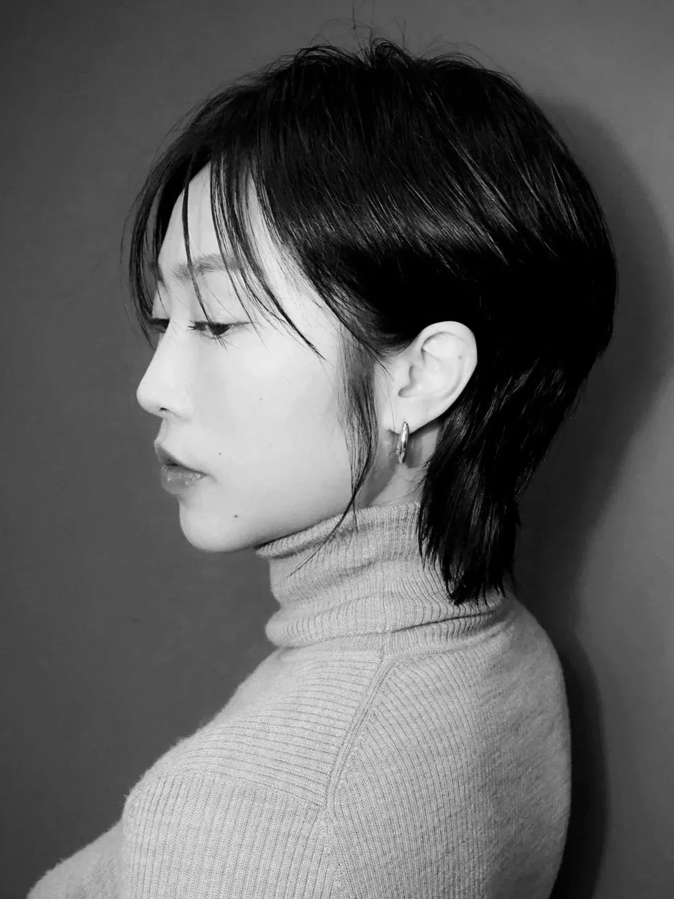 Black and white side-profile portrait of a woman with short, layered hair wearing a turtleneck sweater and hoop earring.