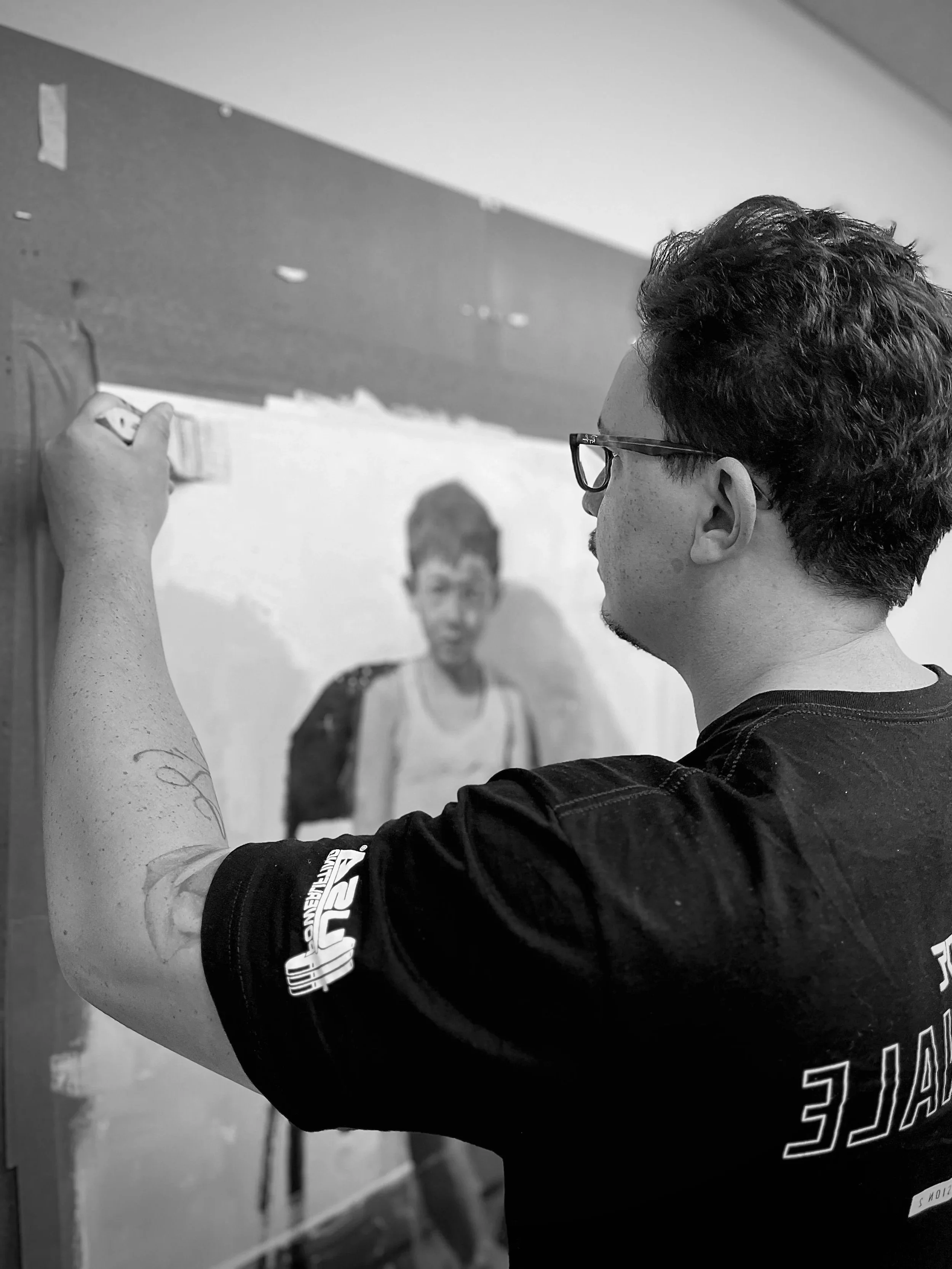 A side view of a man wearing glasses and a black T-shirt as he works on an oil painting of a boy.