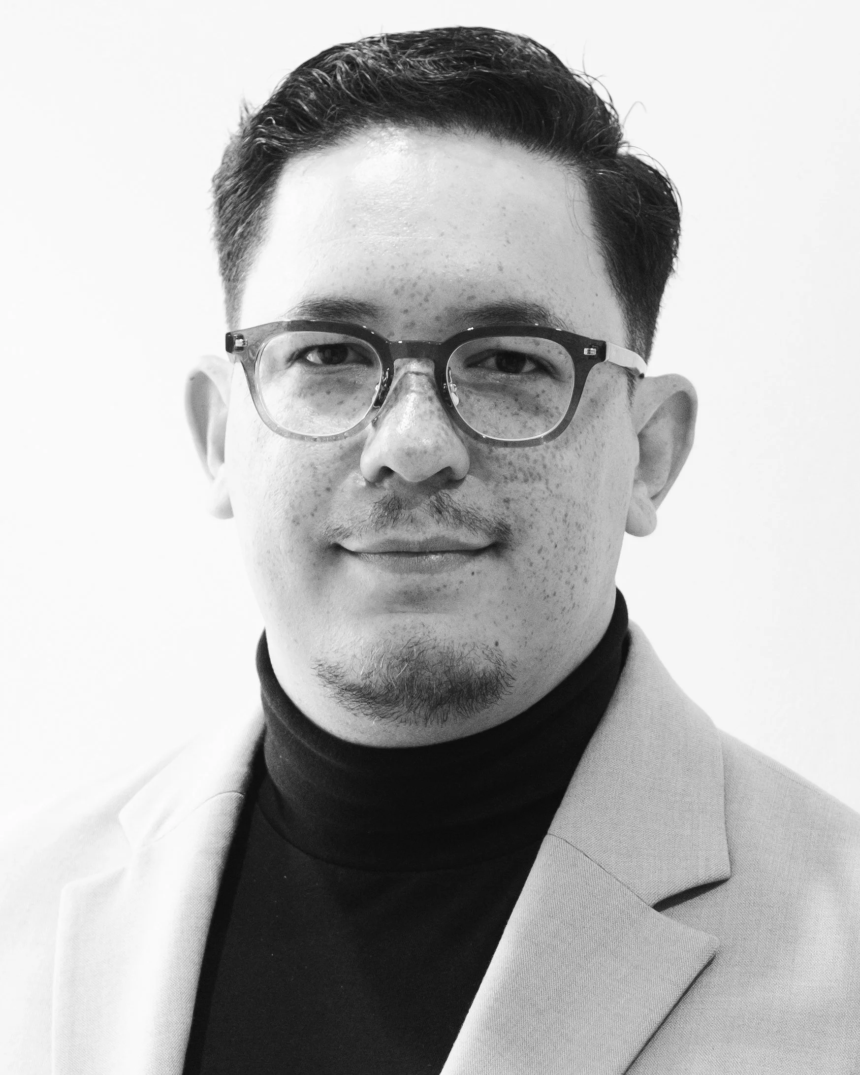 A black-and-white portrait of a young man wearing glasses, a turtleneck, and a blazer, looking at the camera with a slight smile.