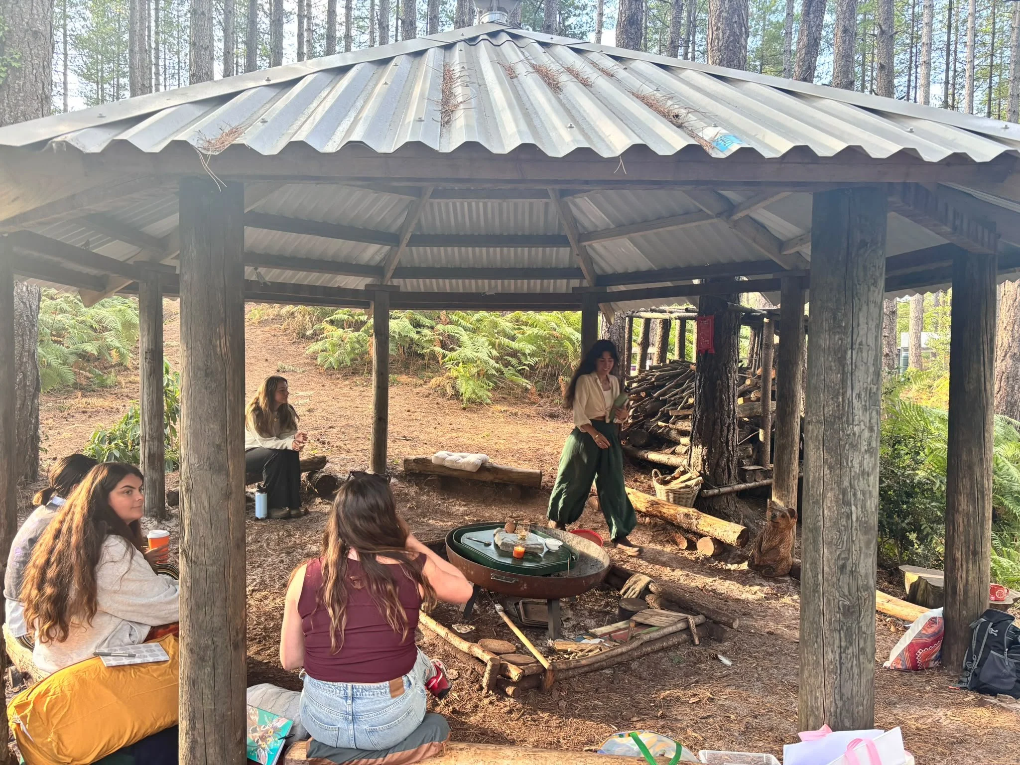 People gathered around a fire pit under a wooden shelter in a forest, some sitting on logs, enjoying an outdoor gathering.