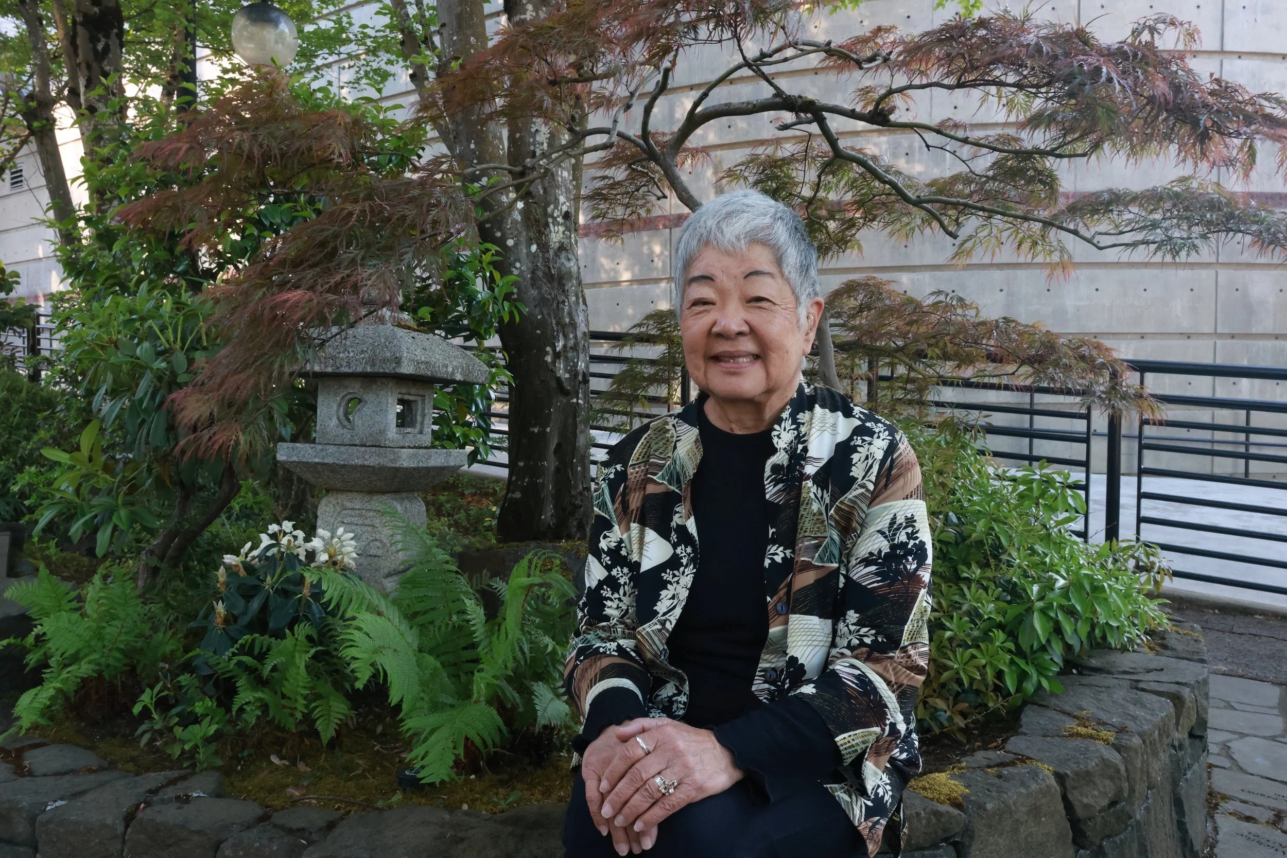 An elderly woman with short gray hair seated in front of a garden with lush green plants, a Japanese stone lantern, a tree with red-brown leaves, and a concrete wall behind her.