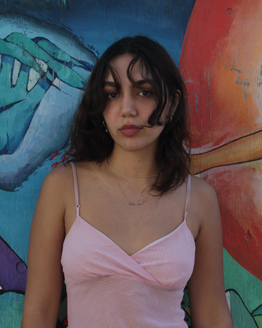 Young woman with shoulder-length dark hair and a pink spaghetti strap dress, standing in front of a colorful abstract mural.