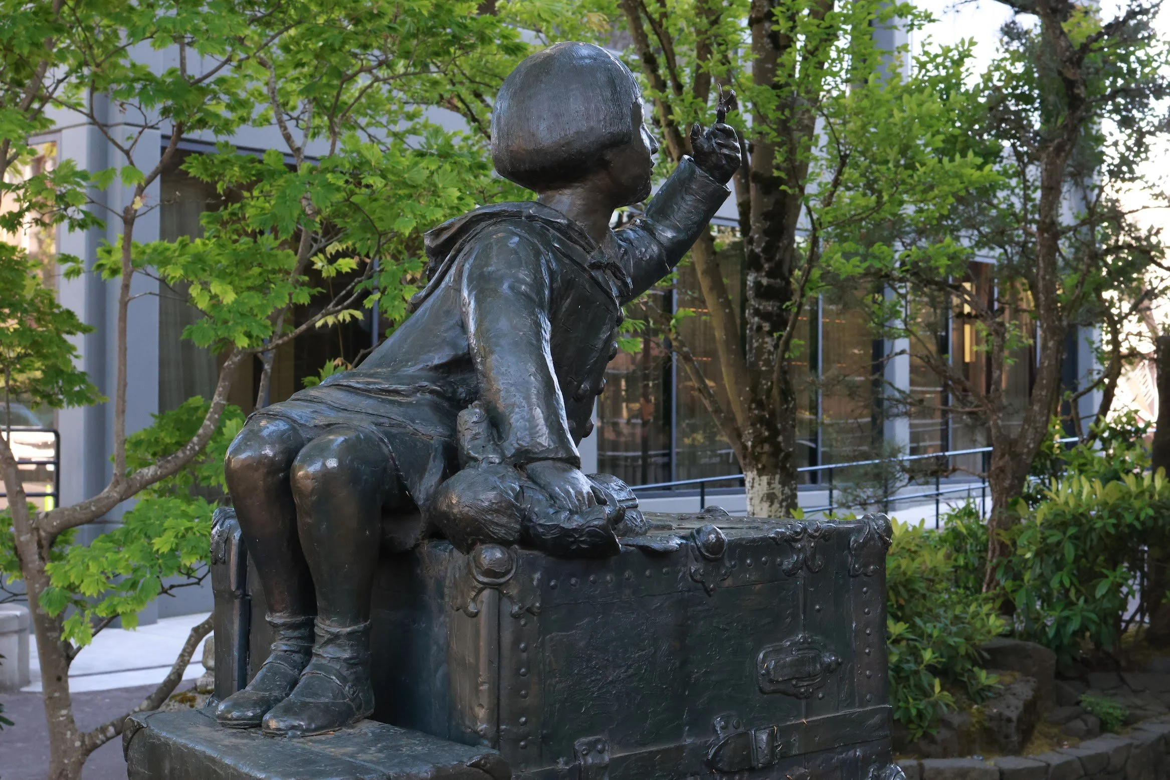 Bronze statue of a young child with a suitcase, sitting on it and raising one hand, surrounded by green trees.