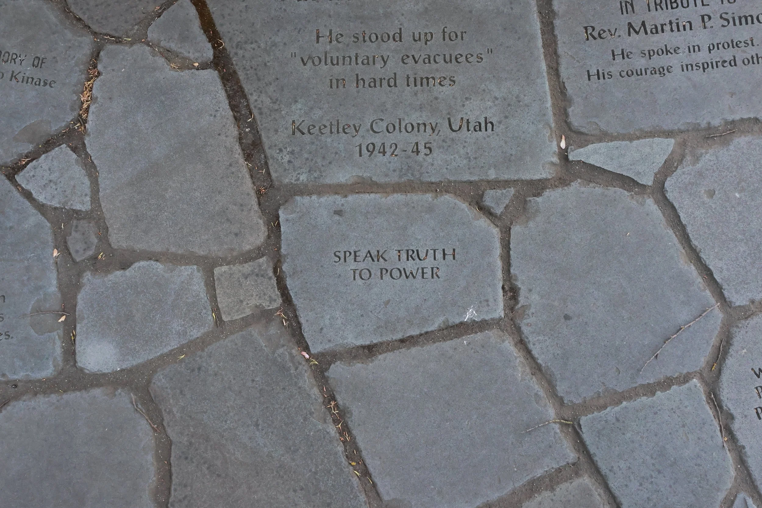 Memorial engraved on a stone in a cobblestone walkway, dedicated to Rev. Martin P. Simo, with inscriptions about speaking in protest and inspired courage, mentioning Keetley Colony, Utah, 1942-45, and the phrase 'Speak Truth to Power'