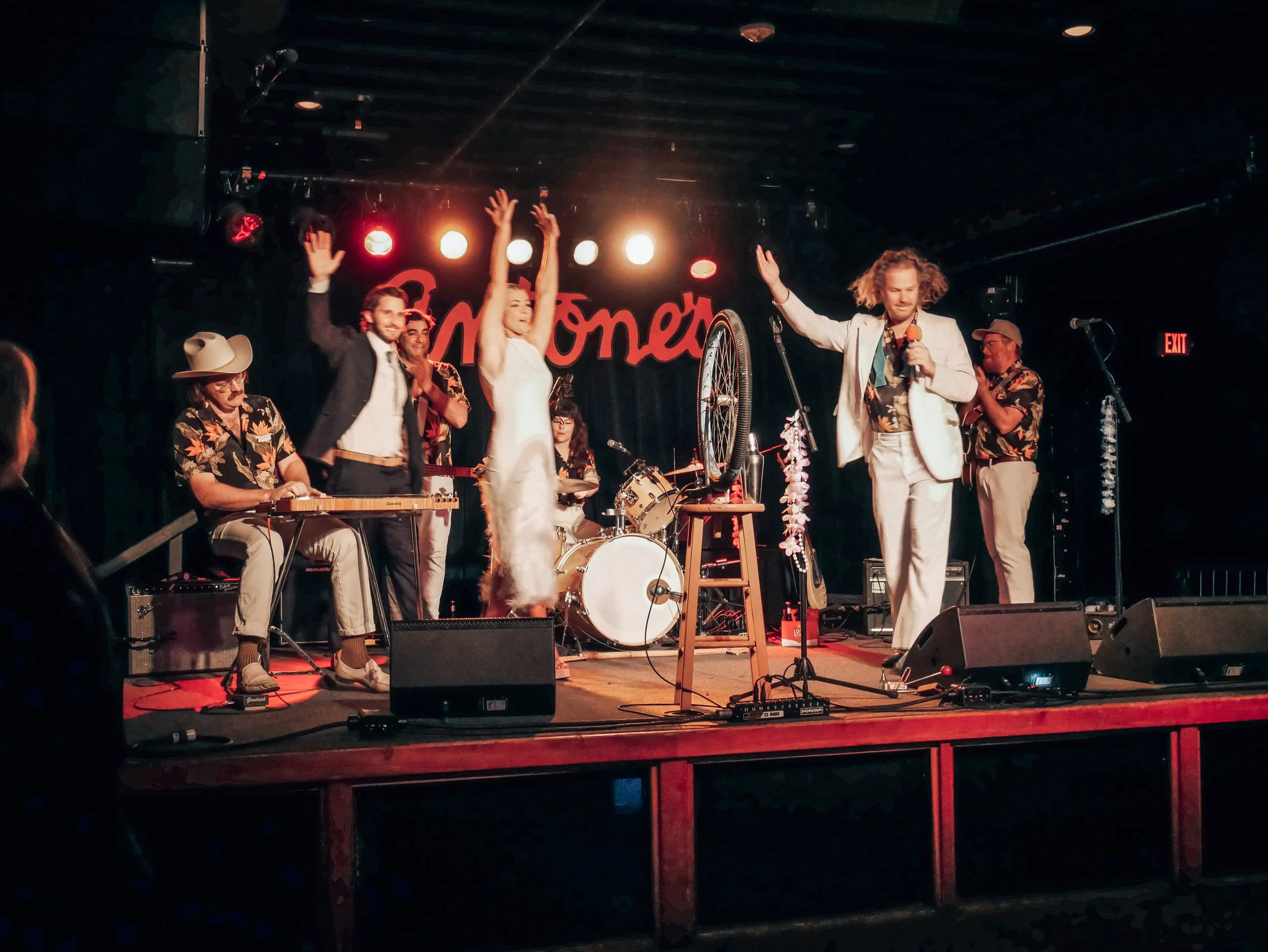 A band performing on stage with a singer holding a microphone, a bride on stage with her arms raised followed by her groom, a guitarist wearing a hat, and other band members, all under red, bright stage lights.