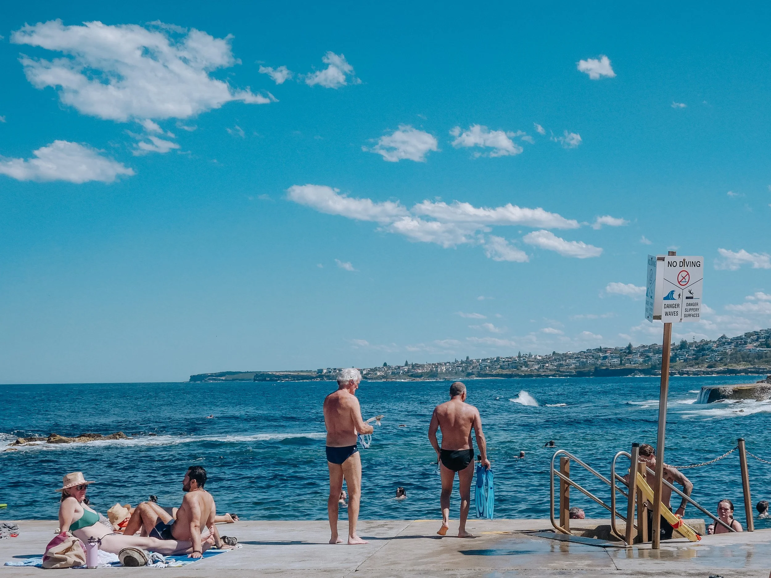 People are enjoying a sunny day at the beach in Australia, some sitting and some preparing to swim, with clear blue sky and ocean waves in the background.