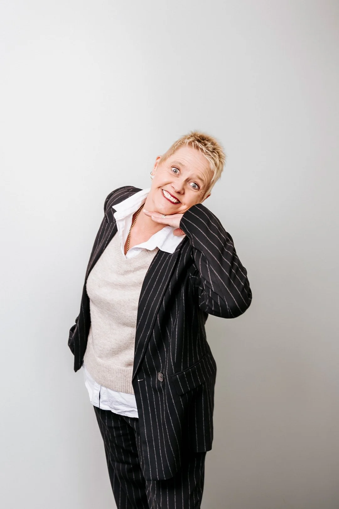 A woman with short blonde hair, wearing a black pinstripe suit over a white shirt and beige vest, smiling and posing with her hand under her chin against a plain light gray background.