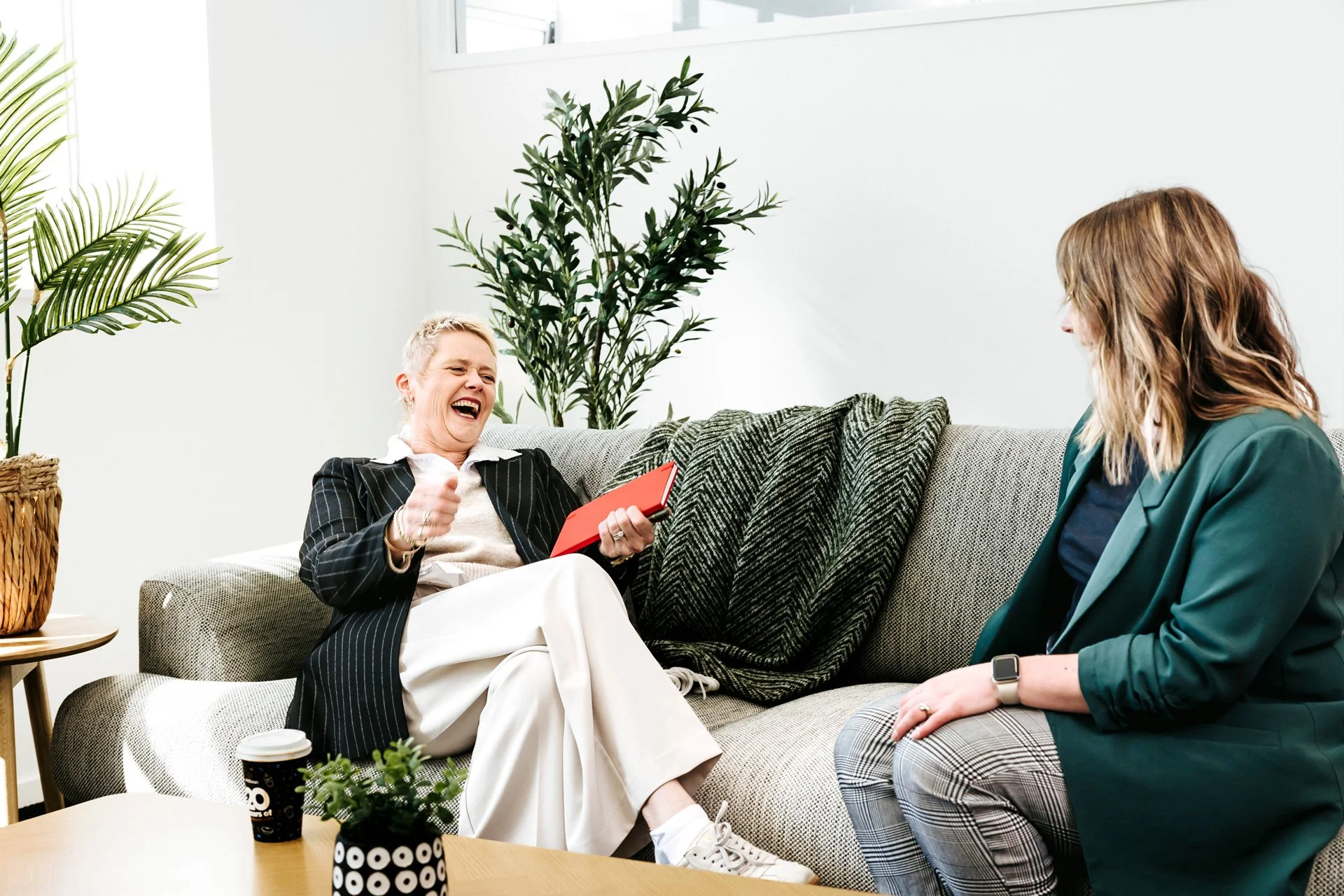 Two women laughing and having a conversation in a modern living room with green plants.