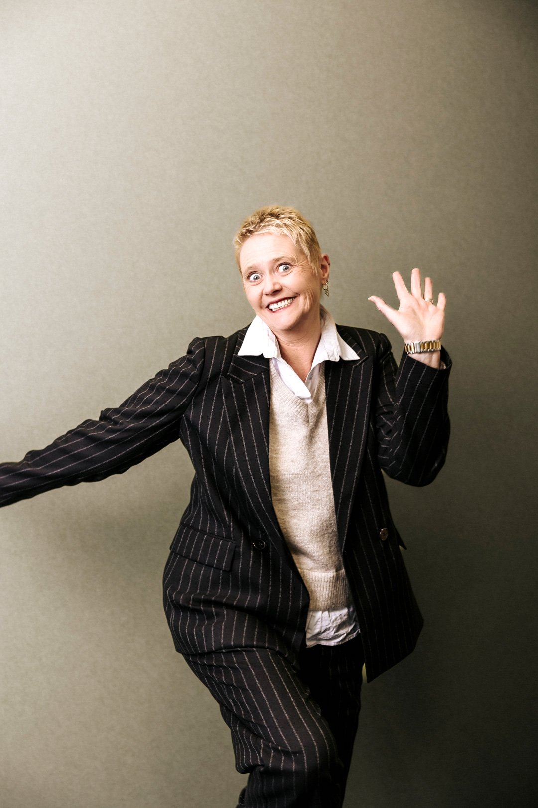 A woman in a black pinstripe suit with a white shirt and beige sweater, smiling and waving against a plain green background.
