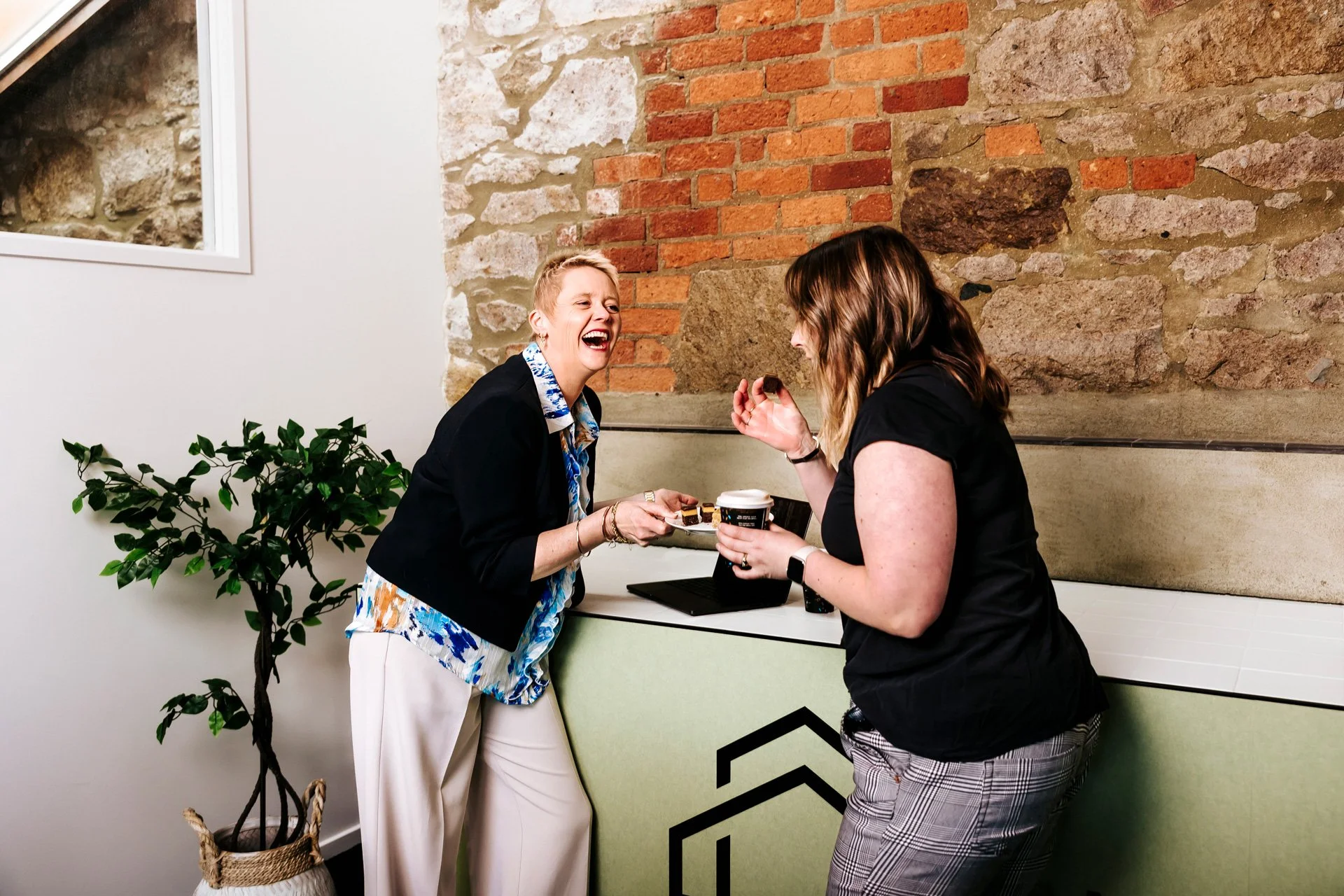 Two women are laughing and enjoying a moment together at a cafe, one holding a piece of cake and the other holding a coffee cup, standing near a brick and stone wall behind a counter with a plant nearby.