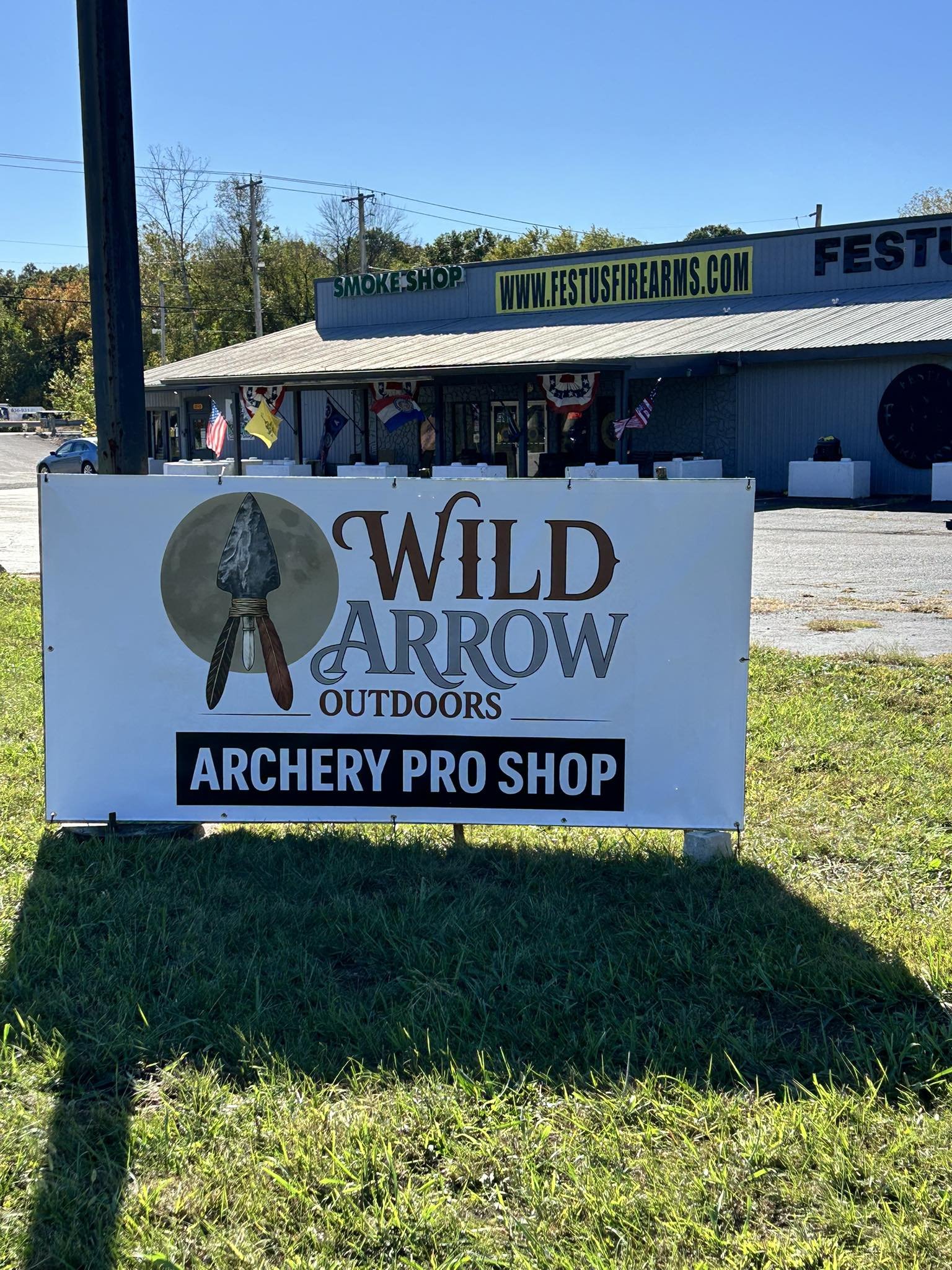 Sign for Wild Arrow Outdoors Archery Pro Shop in front of a building with a large sign, flags, and outdoor decorations.