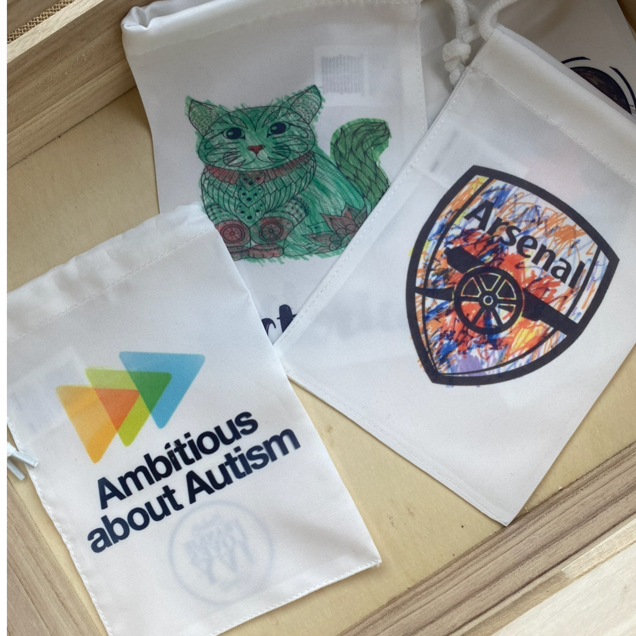 Three white fabric drawstring pouches with colorful logos and designs, including a green and red cat illustration, a 'Arsenal' logo with a cannon, and a message about autism awareness.