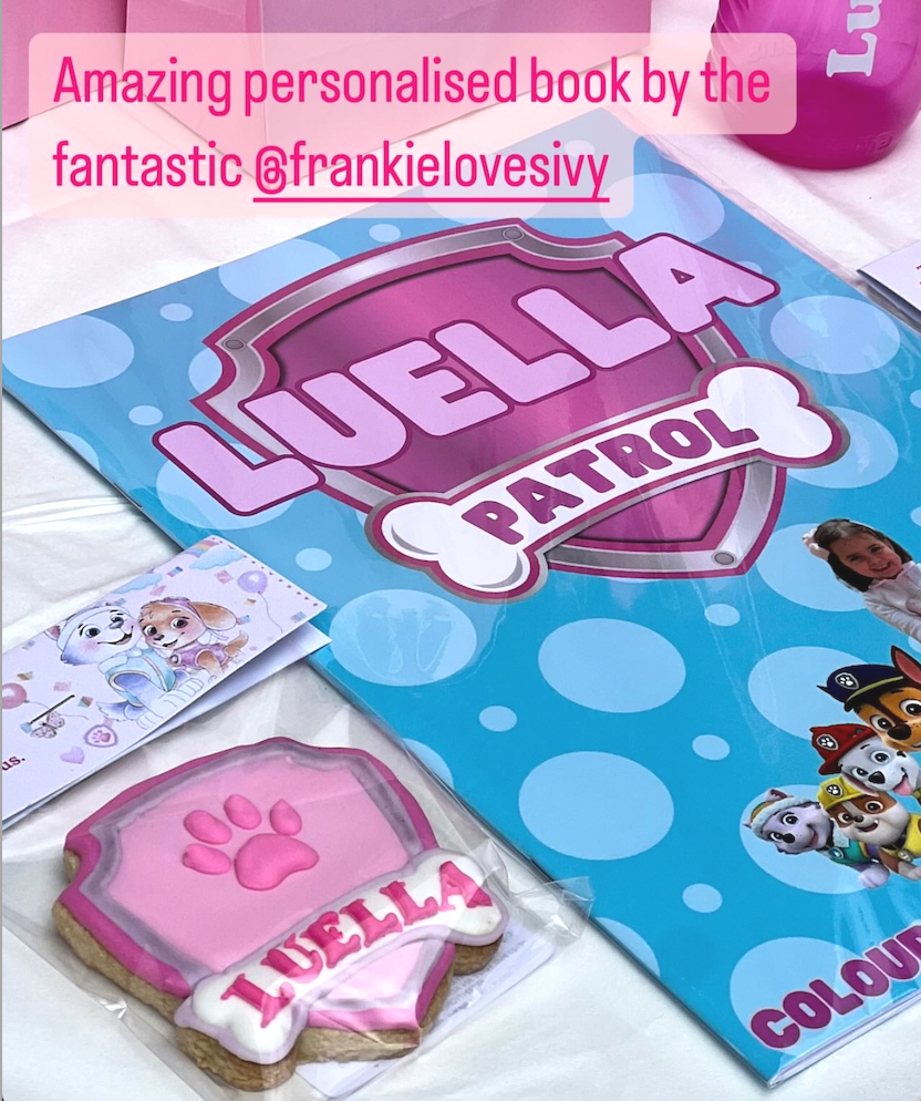 A colorful children's book titled 'Paw Patrol' with a large badge on the cover, a pink and white paw print cookie with '1 UELLA' written on it, a card featuring characters from Paw Patrol, and a pink mug in the upper right corner.