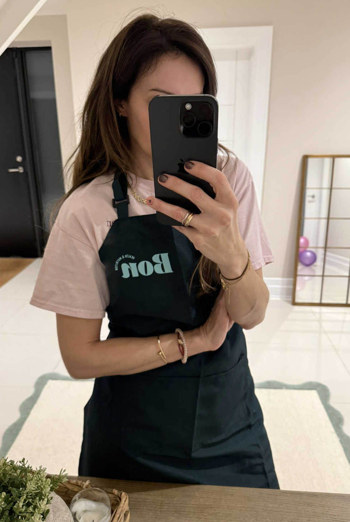 Woman taking a mirror selfie wearing a pink t-shirt and a black apron with teal text that says 'Bon'. She has long brown hair and jewelry on her wrists and fingers, and is standing in a room with a white tile floor, a decorative mirror with balloons in the background, and a plant with a candle on a wooden table in the foreground.
