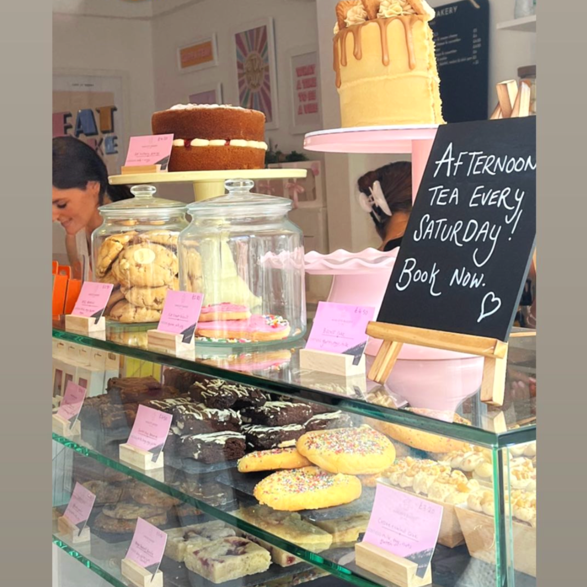Display case with various baked goods including cookies, biscuits, and cakes, with a blackboard sign advertising Saturday afternoon tea and a two-tier cake on a pink stand.
