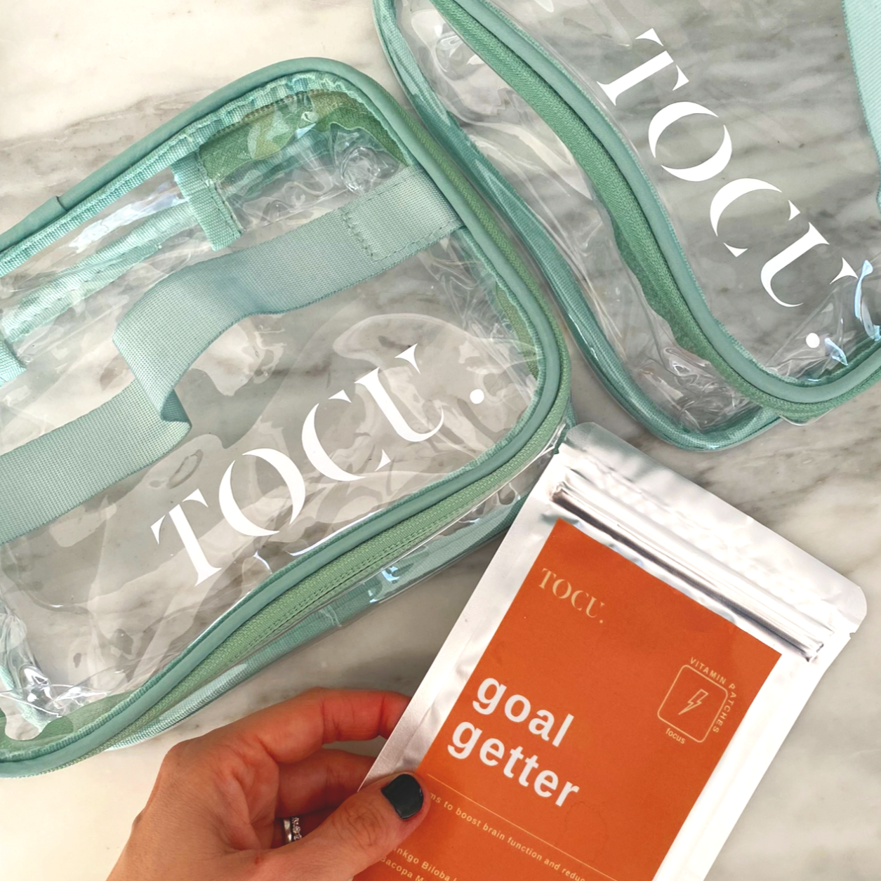 Clear storage bags with teal accents and a package labeled "goal getter" held by a person with black nail polish. The bags are on a marble surface.