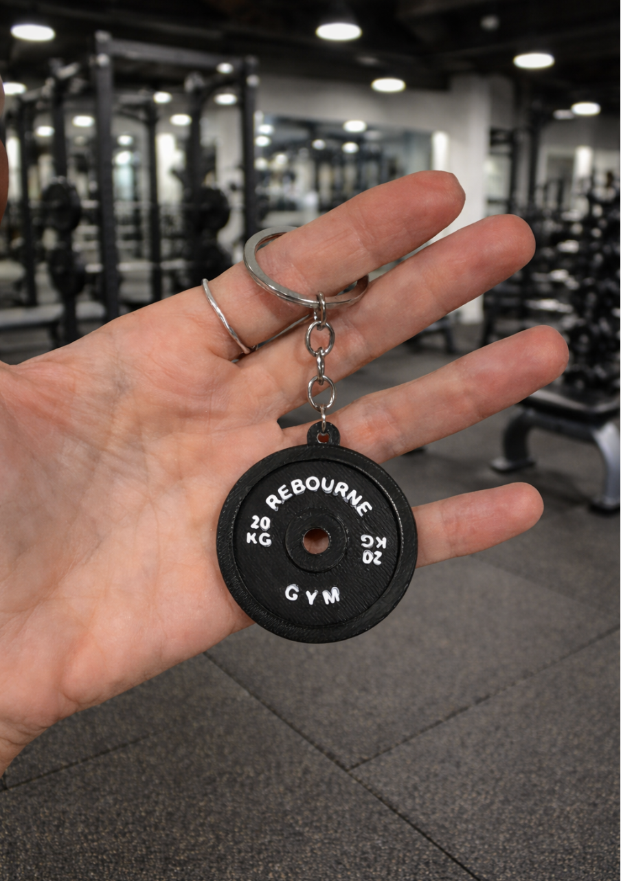 A person's hand holding a gym keychain with a black weight plate design, displaying 'Rebourne Gym' and weight markings, inside a gym with exercise equipment in the background.