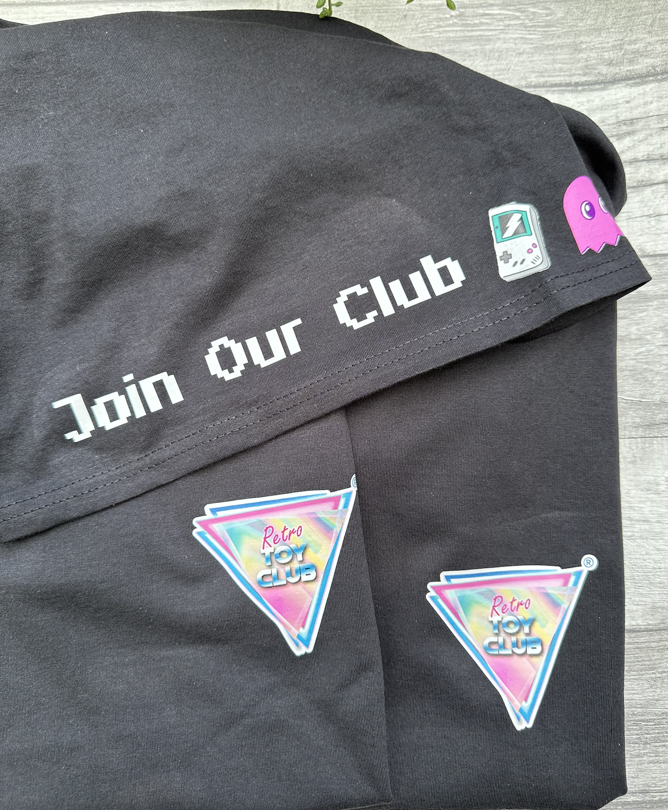 Black fabric with white text that reads 'Join our club', two colorful Retro Toy Club logos, a small vintage gaming console sticker, and a pink ghost sticker, all on a wooden surface.