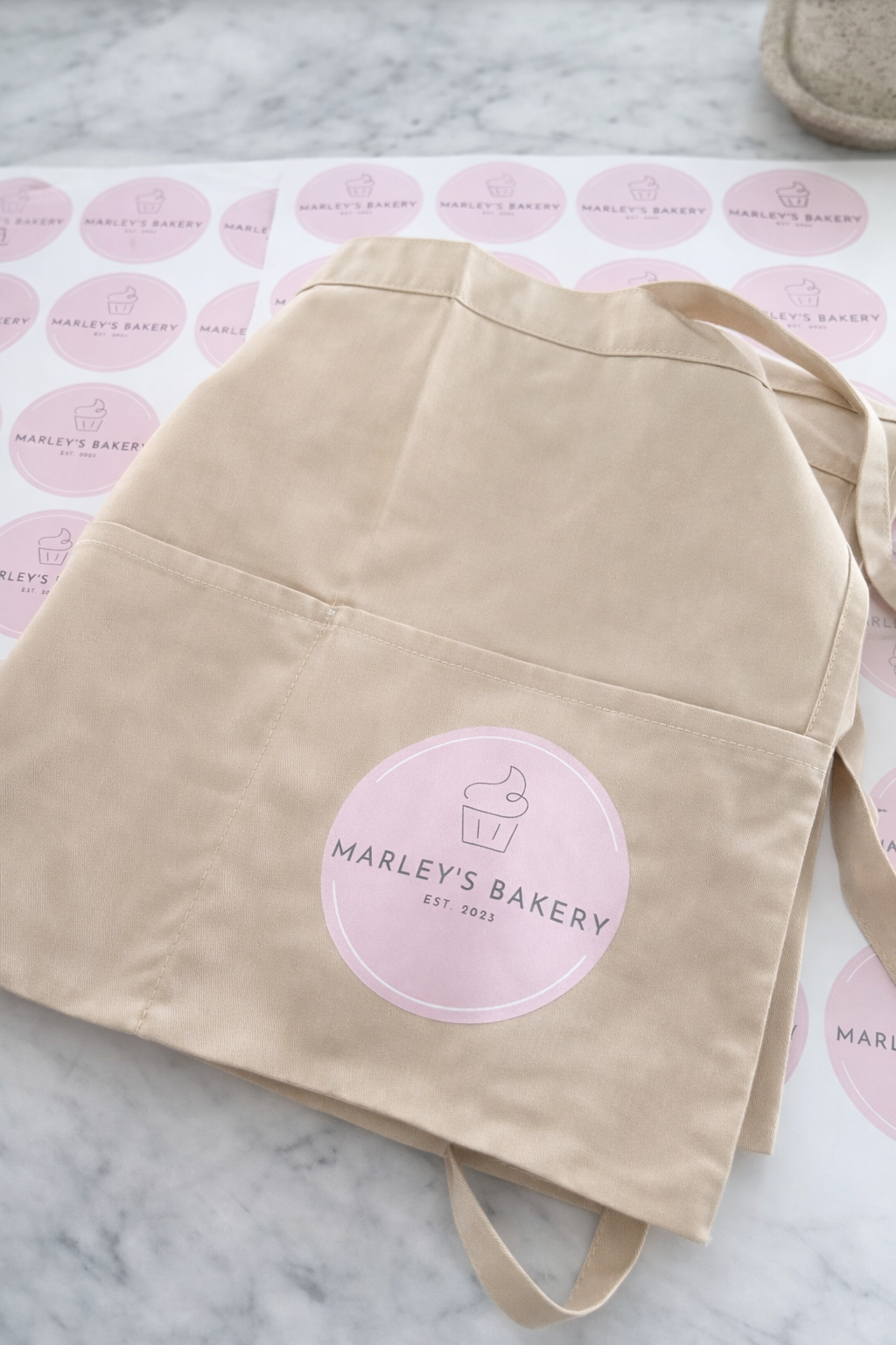 A beige tote bag with the pink logo of 'Marley's Bakery, EST. 2023' on a marble surface with bakery paper underneath.