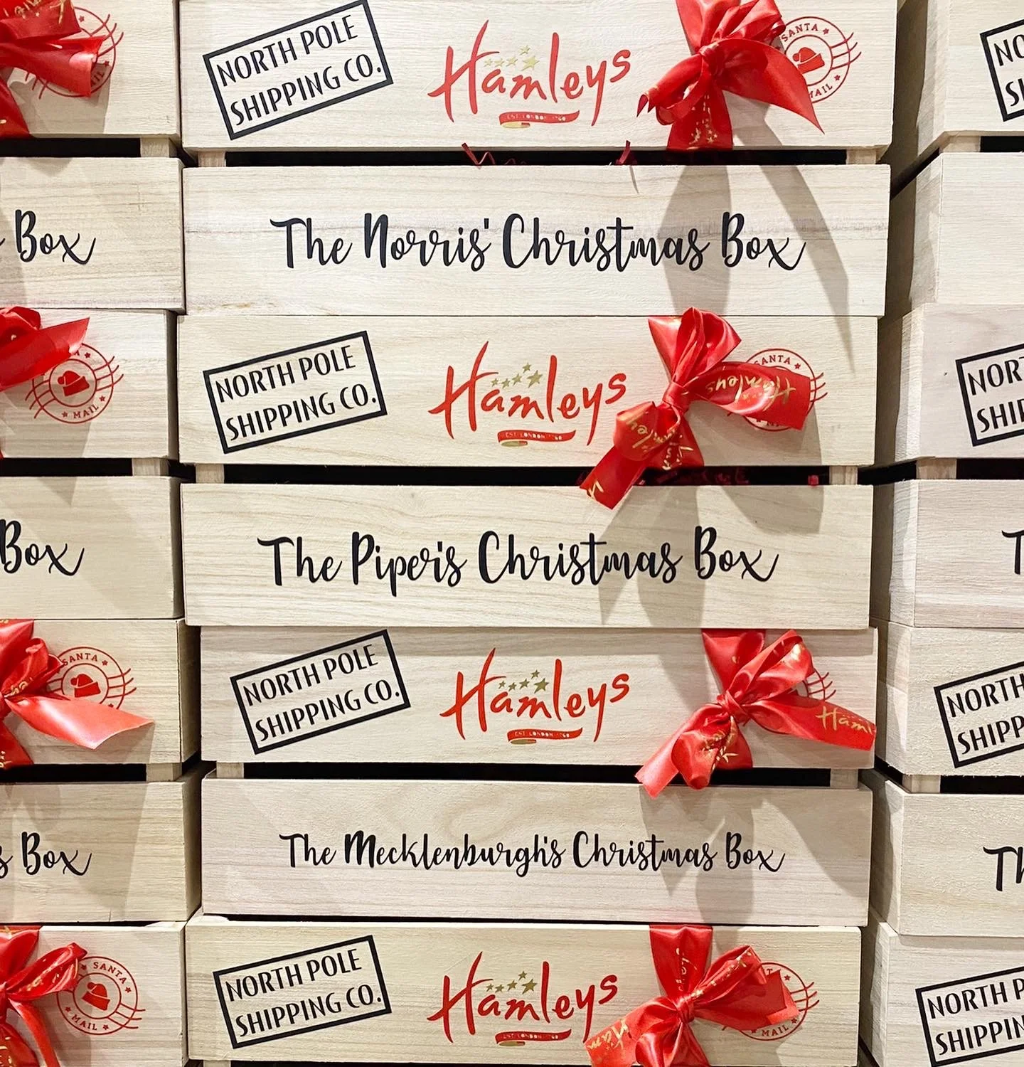 Stacks of Christmas gift boxes labeled with different holiday-themed names, each decorated with a red ribbon, from North Pole Shipping Co. and Hamley's, including 'The Norris' Christmas Box,' 'The Piper's Christmas Box,' and 'The Mecklenburgh's Christmas Box.'