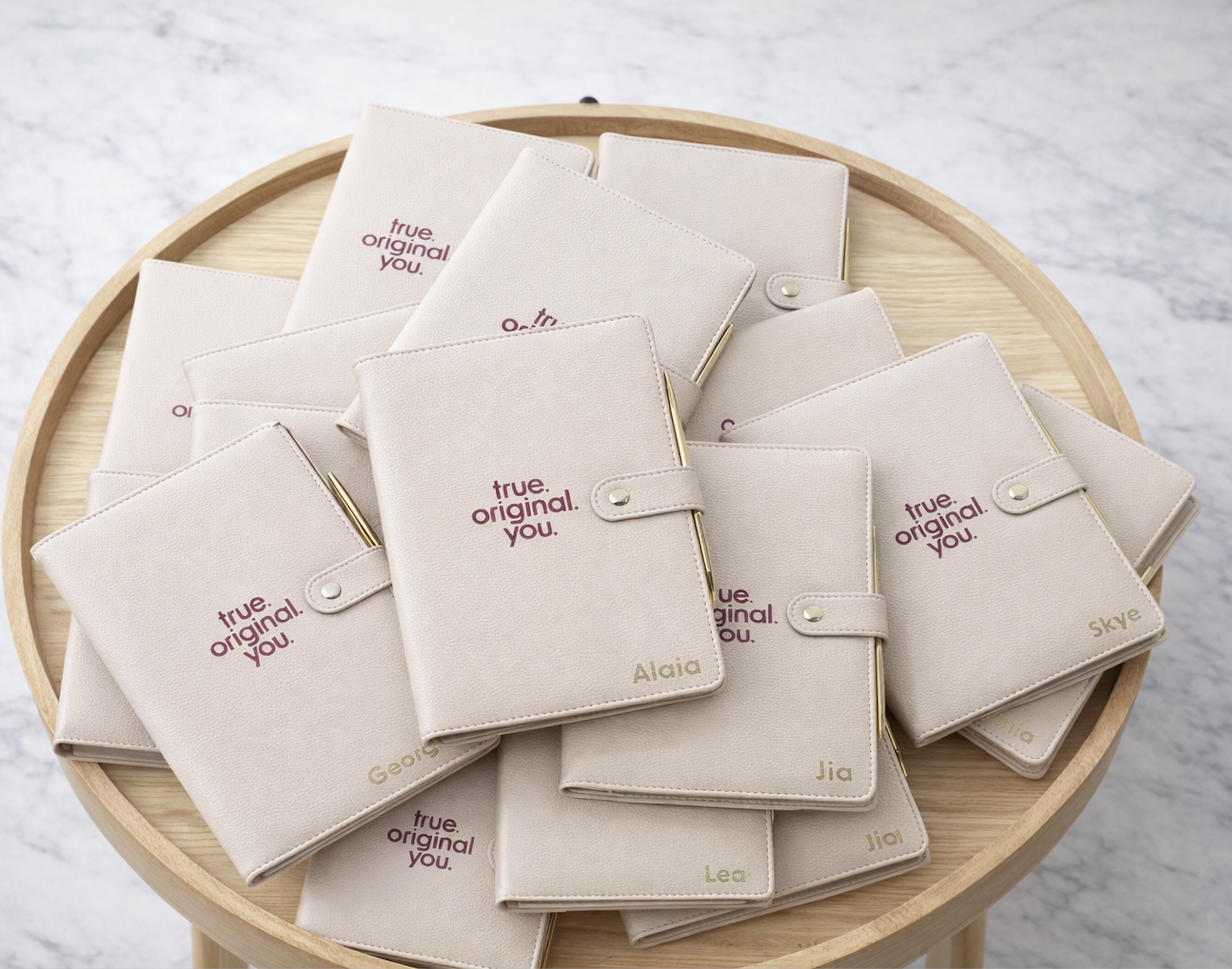 Multiple light beige notebooks with the phrase 'true. original. you.' printed in purple and personalized with gold names, placed on a wooden tray.
