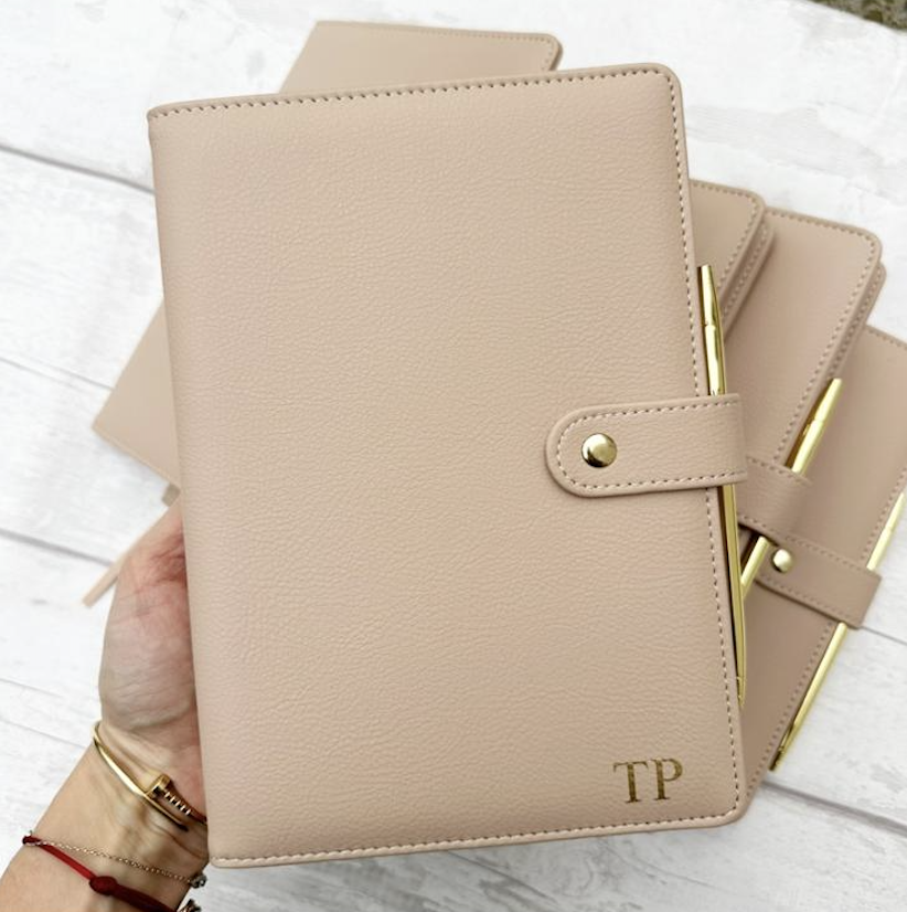 Three beige planners with gold pens and embossed initials "TP" on the cover, held by a hand with bracelets, on a white wooden surface.