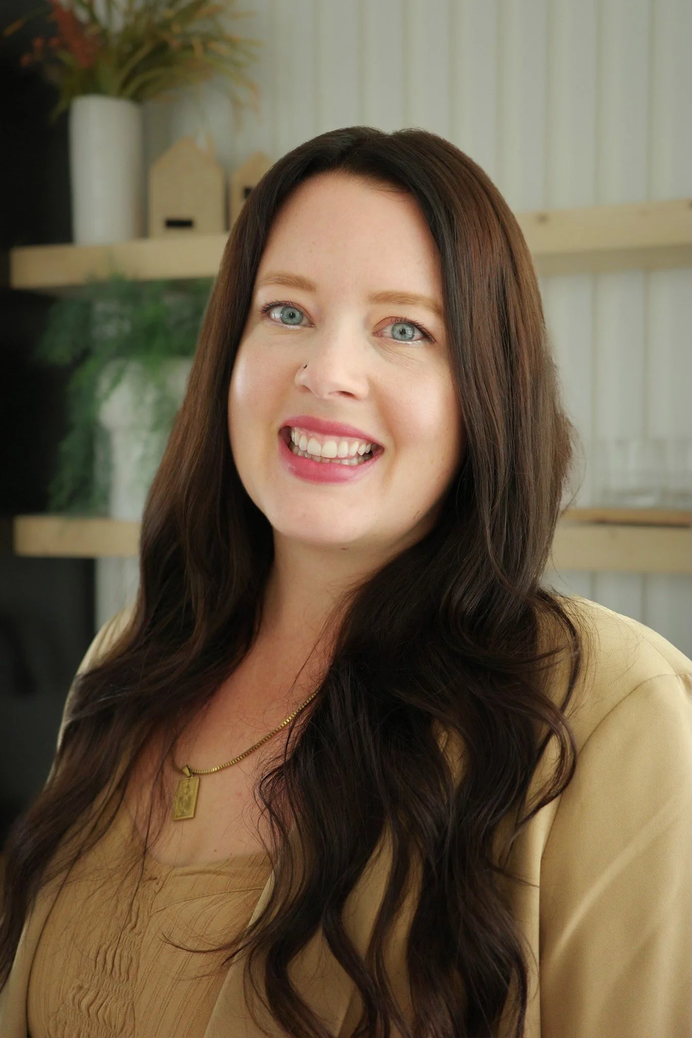 A woman with long dark brown hair, blue eyes, and fair skin smiling at the camera, wearing a beige top and a gold necklace, standing indoors with a bookshelf and decorative objects in the background.