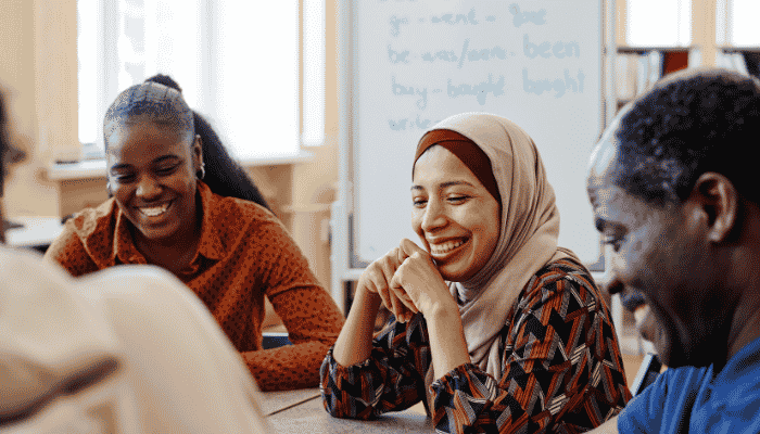 Three diverse women sitting at a table in a classroom, smiling and talking.