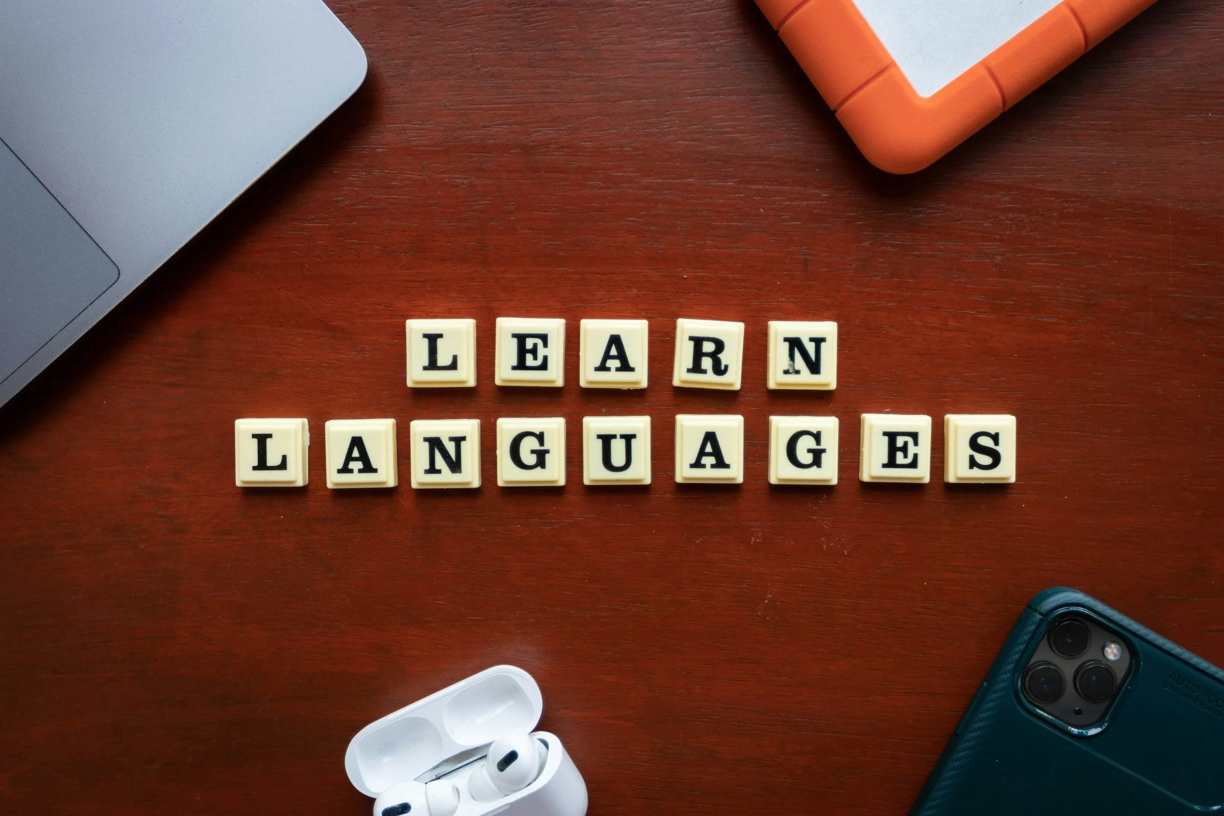 Scrabble tiles spelling 'LEARN LANGUAGES' on a wooden desk with a laptop, orange case, and wireless earbuds nearby.
