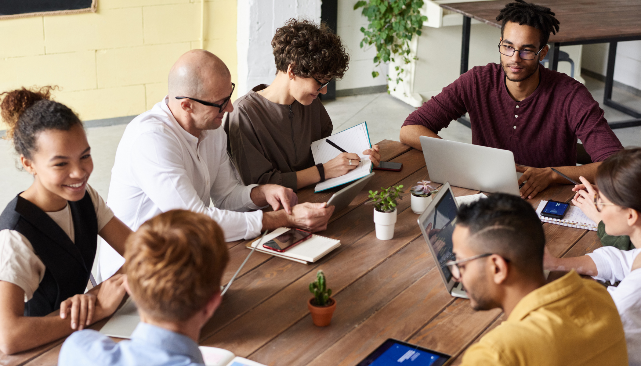 A diverse group of people sitting around a wooden table, engaged in a discussion with laptops, tablets, notebooks, and smartphones, in a well-lit room with yellow walls and plants.