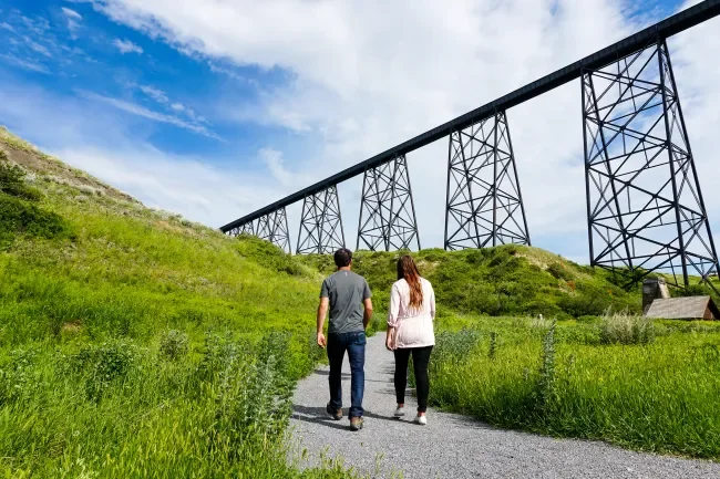 A couple walking on a gravel path through green grass with a large steel train trestle bridge in the background under a partly cloudy sky.
