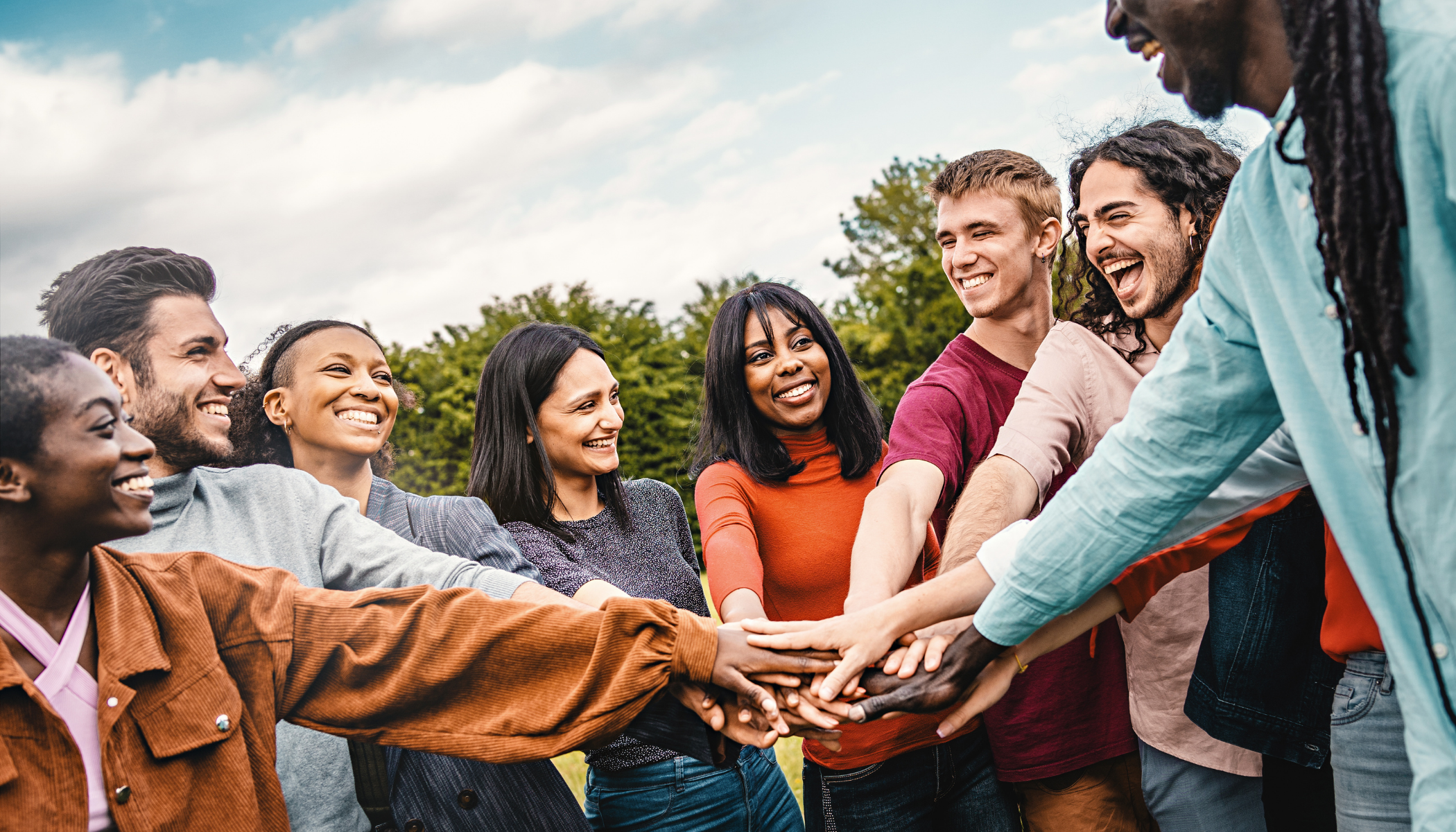 Group of diverse smiling people with hands together in an outdoor setting, celebrating teamwork or unity.